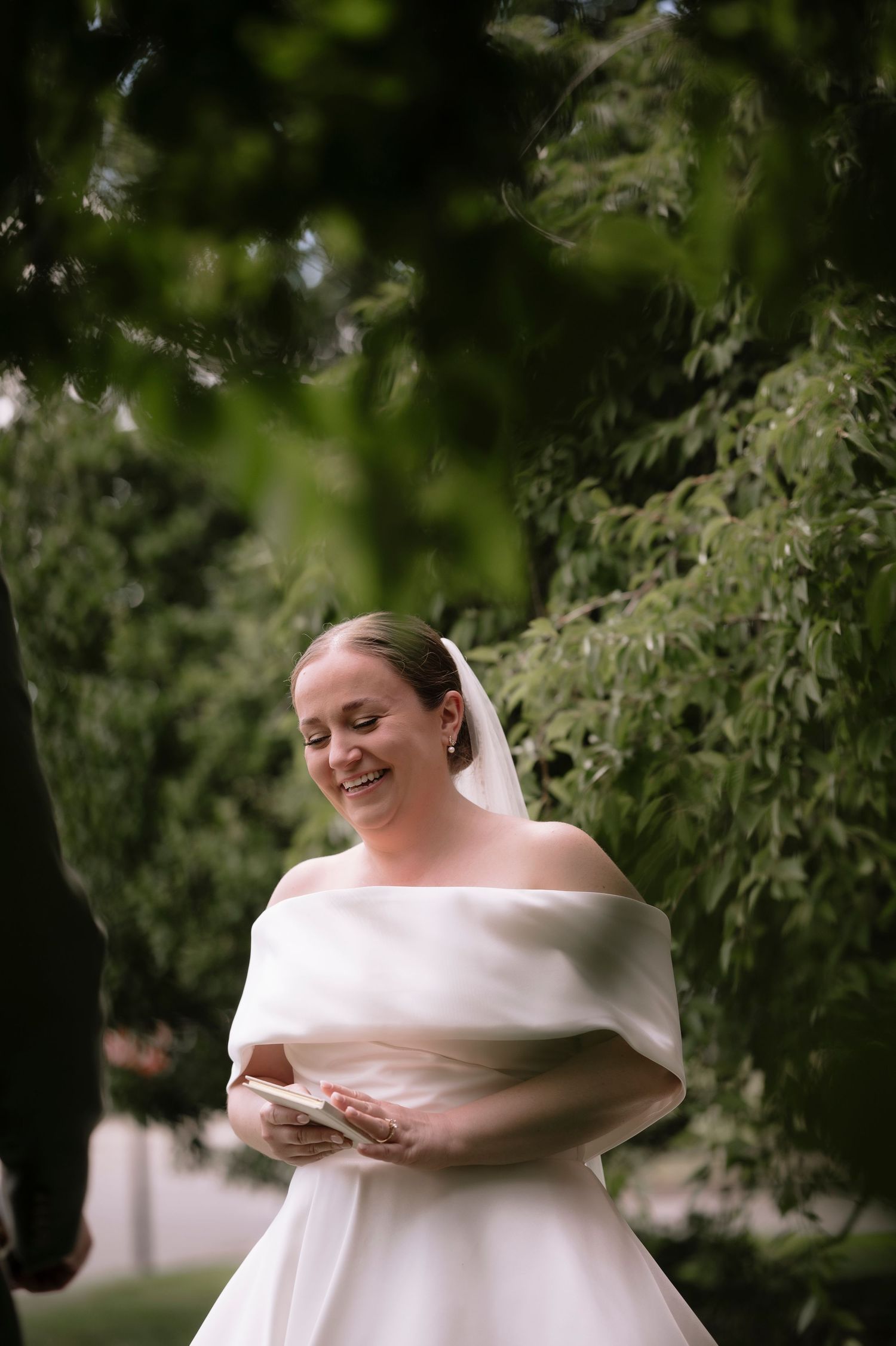 Smiling bride in off-shoulder white gown and veil reads note outdoors among lush green trees.