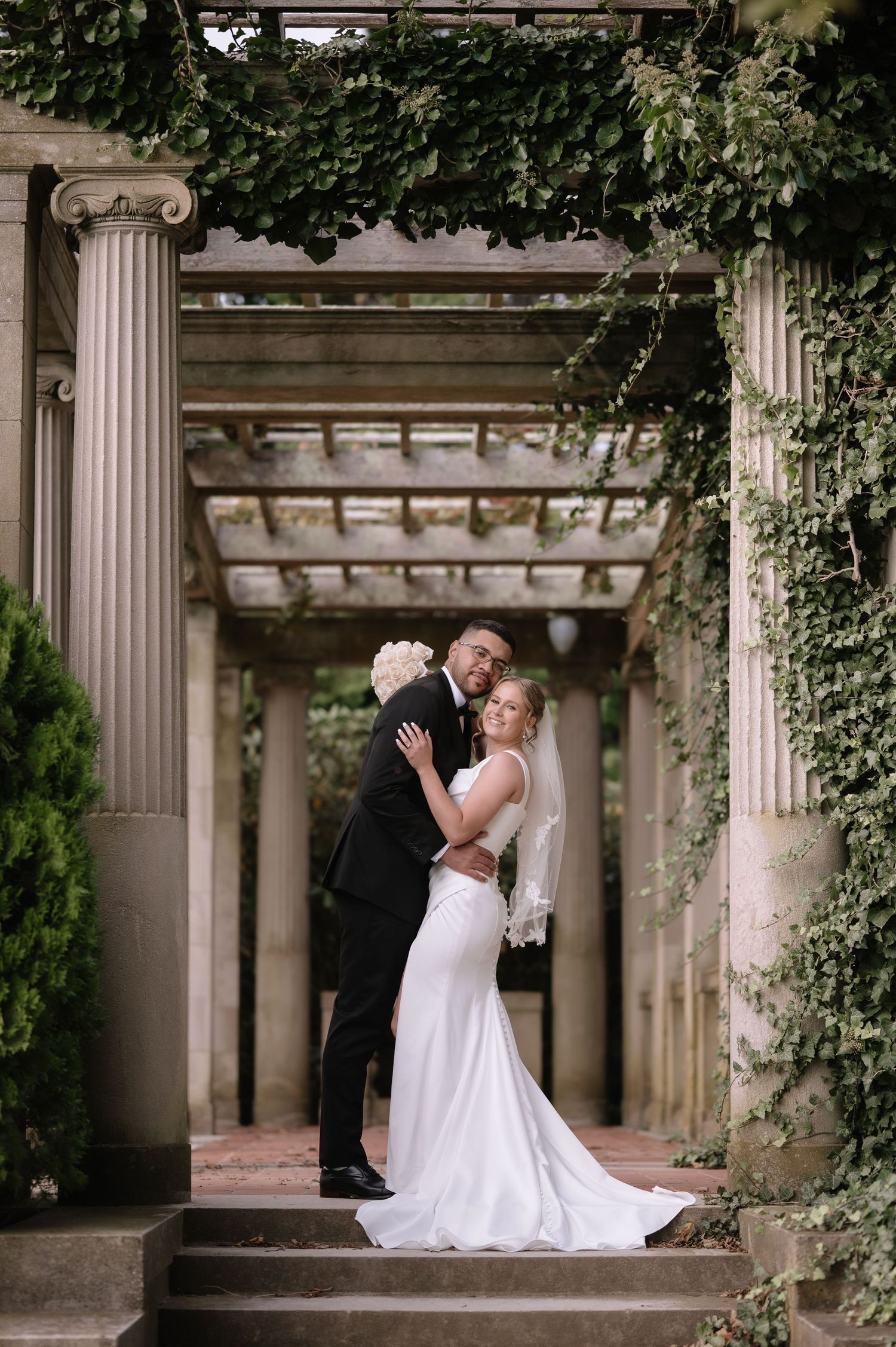 Romantic wedding couple poses in elegant colonnade setting with ivy-covered pillars and classical architecture.