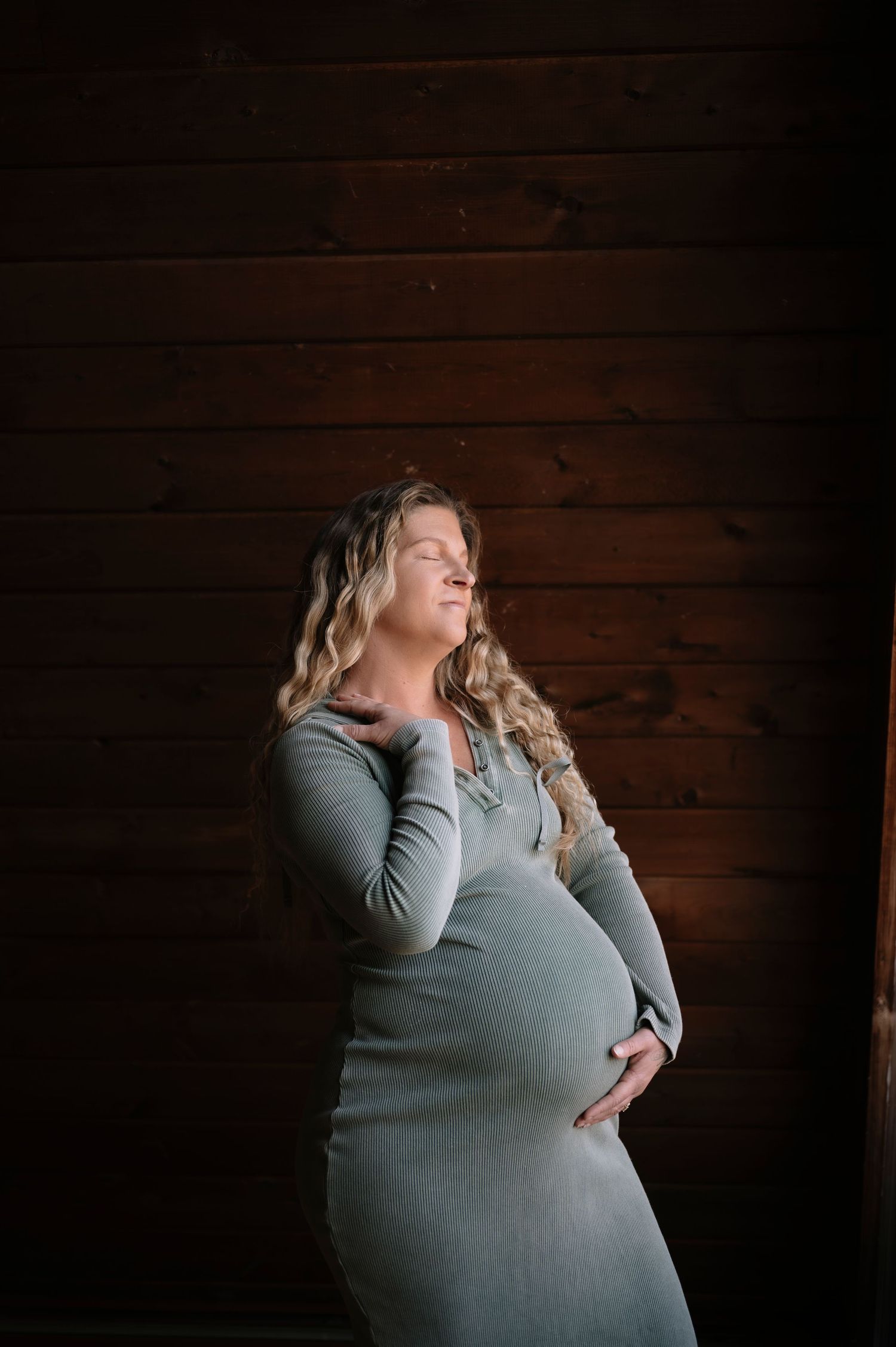 A pregnant woman in a gray sweater poses against a dark wooden wall in a series of artistic maternity photos.
