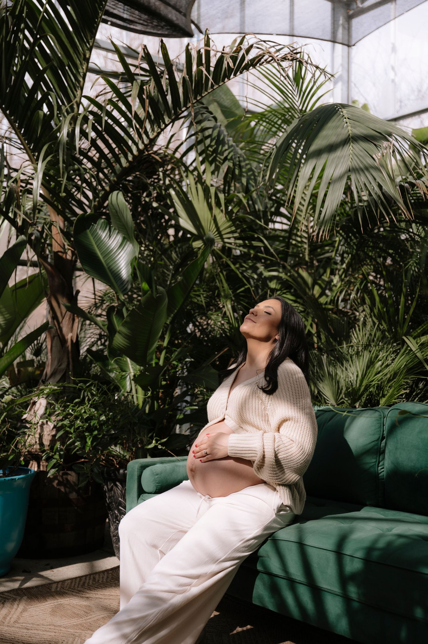 A pregnant woman in a cream sweater relaxes on a green couch surrounded by lush tropical plants in a sunlit conservatory.