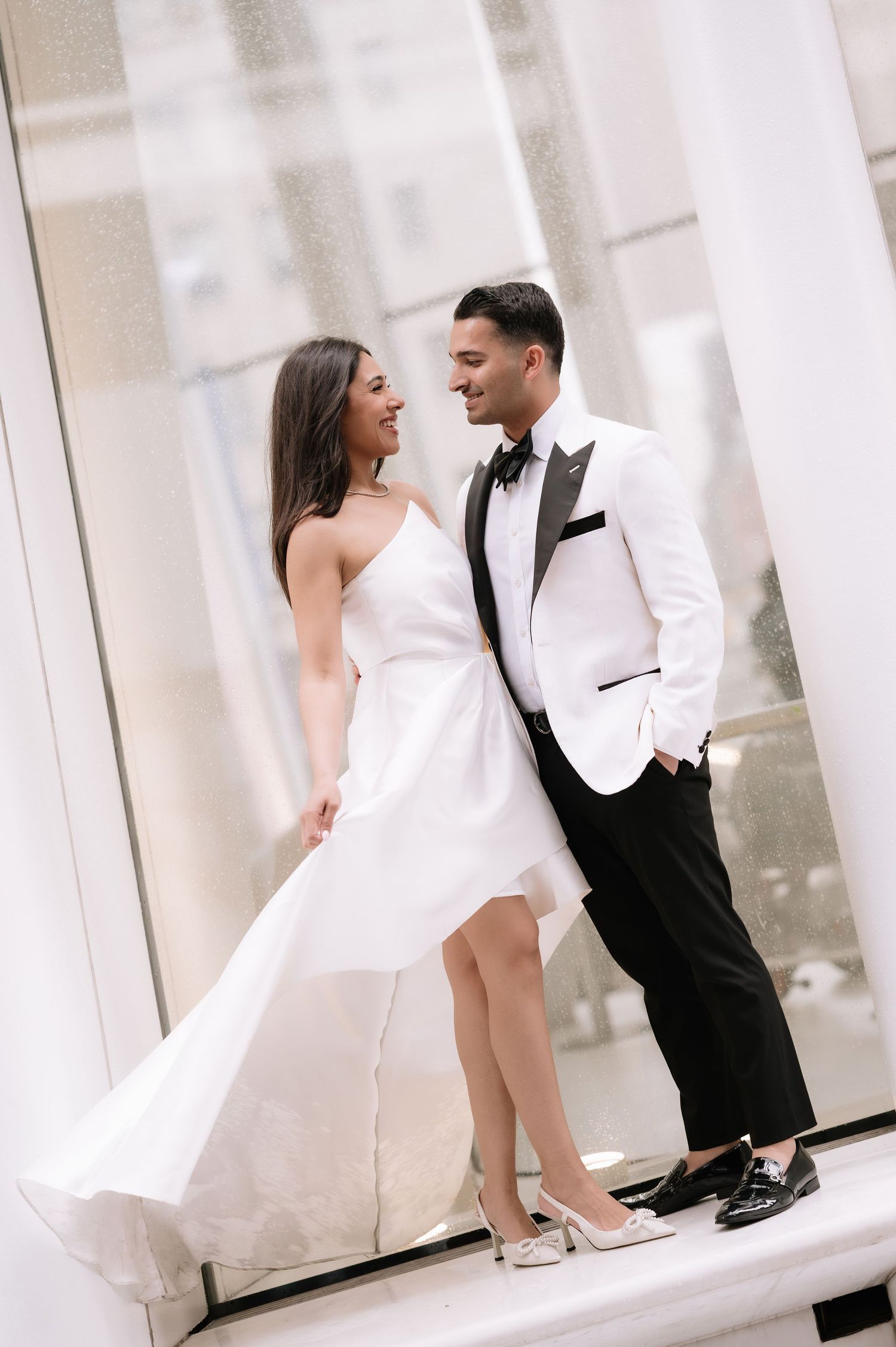 Romantic couple in wedding attire laughing together against white pillars in modern setting.