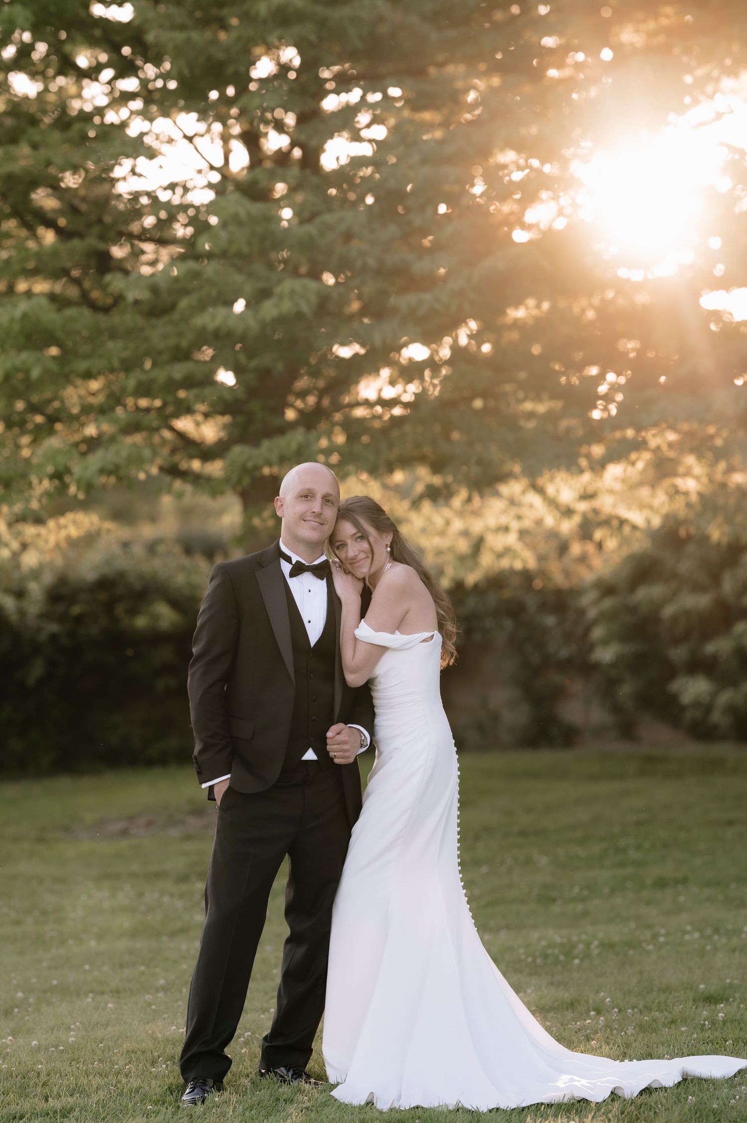 A couple poses outdoors at sunset for their wedding photo with golden light filtering through trees behind them.