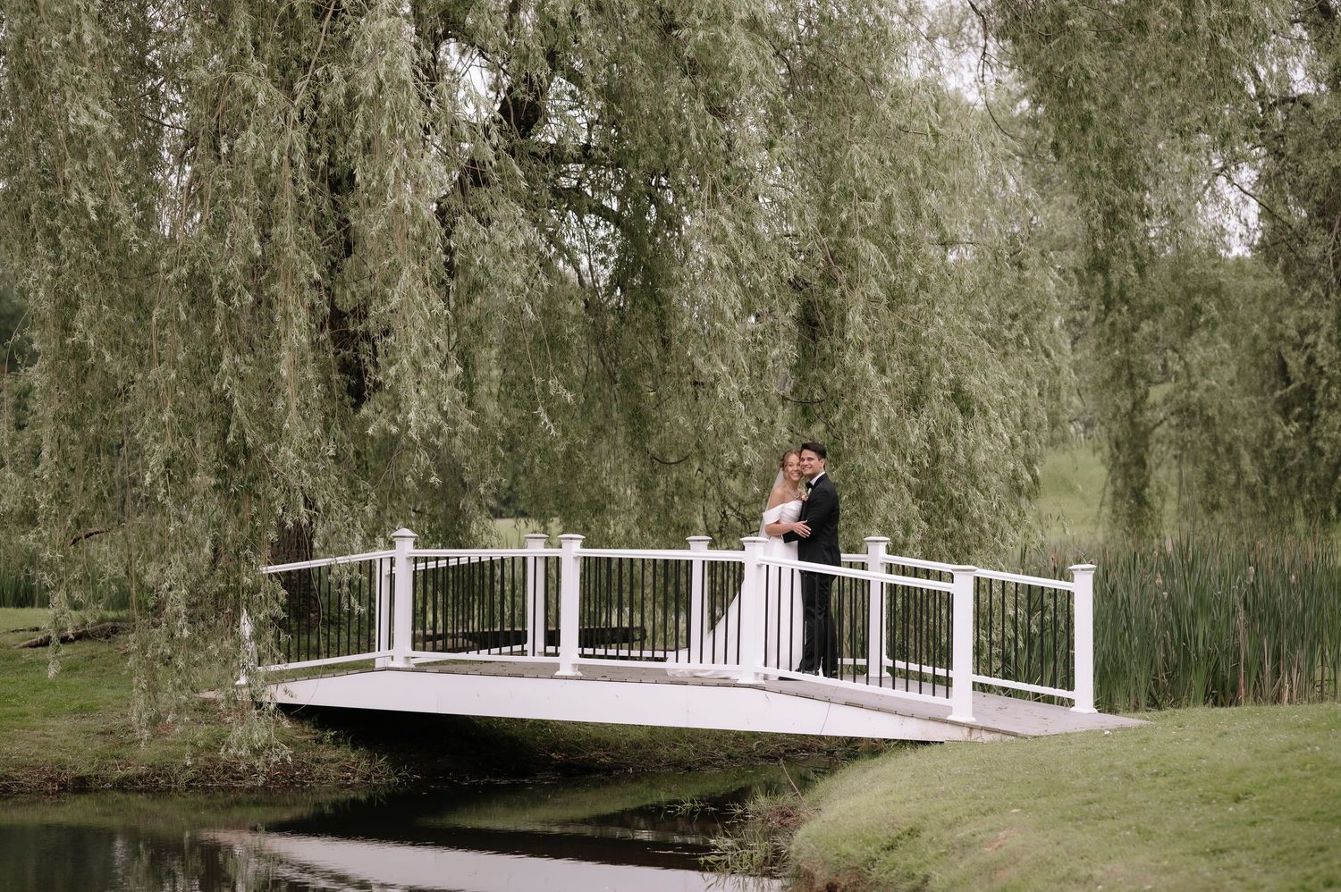 A couple takes a romantic moment on a white arched bridge surrounded by weeping willow trees over a pond.