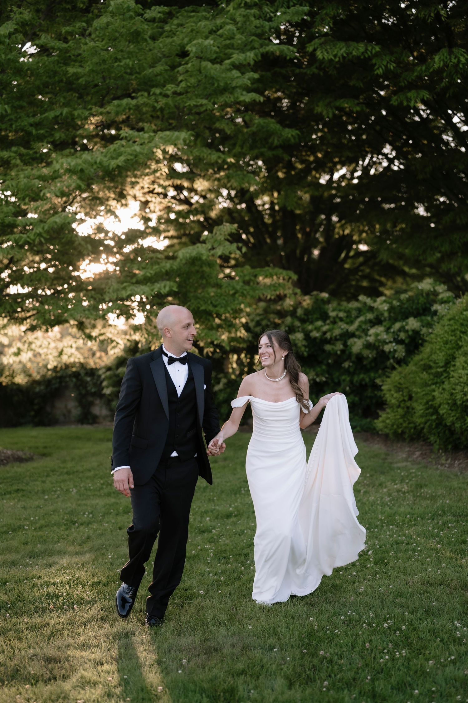 A couple in wedding attire walks hand in hand on a grassy lawn with lush trees and sunset lighting in the background.