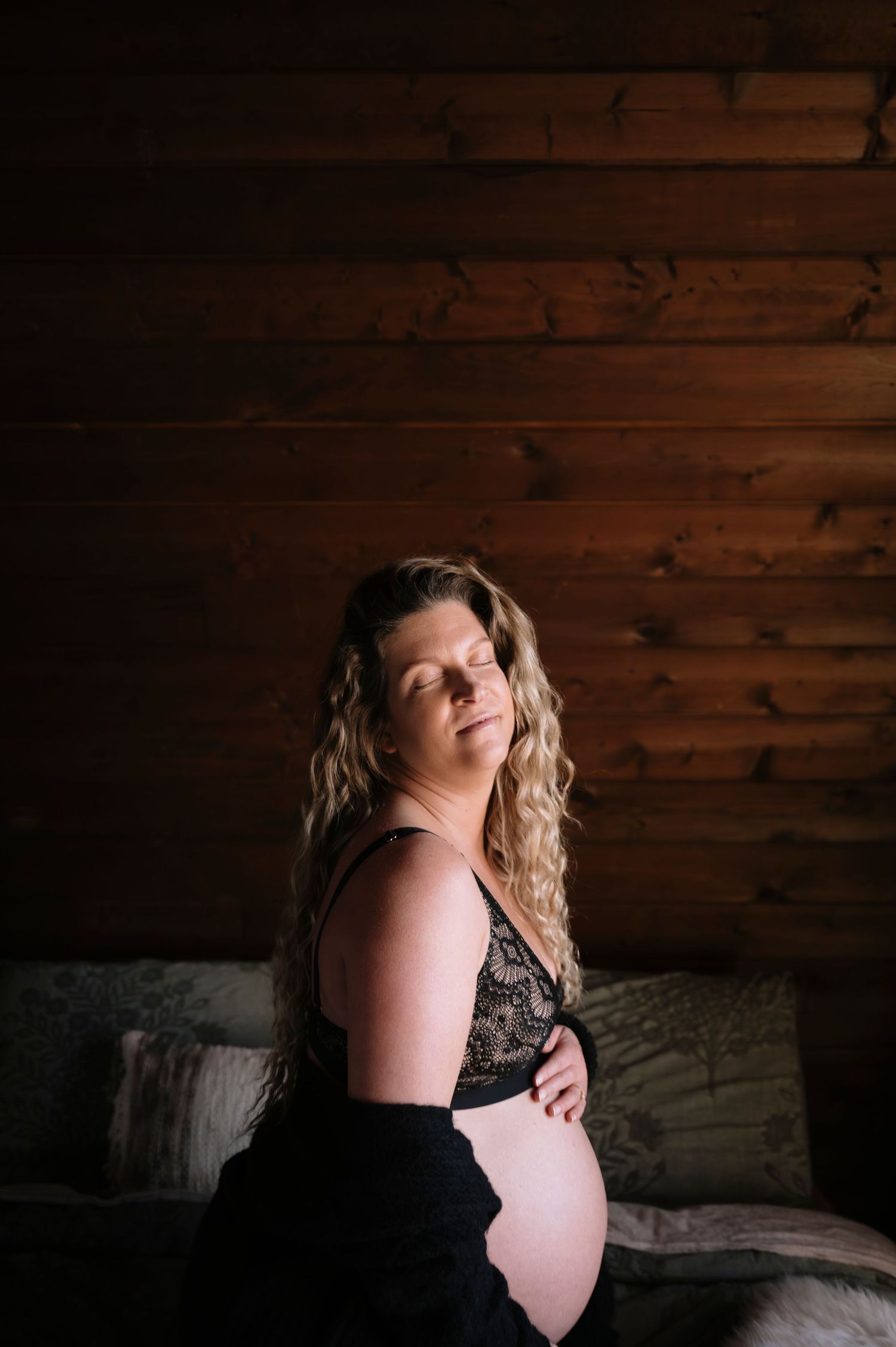 Artistic boudoir photo with moody lighting shows a person in black lingerie against rustic wooden wall backdrop.