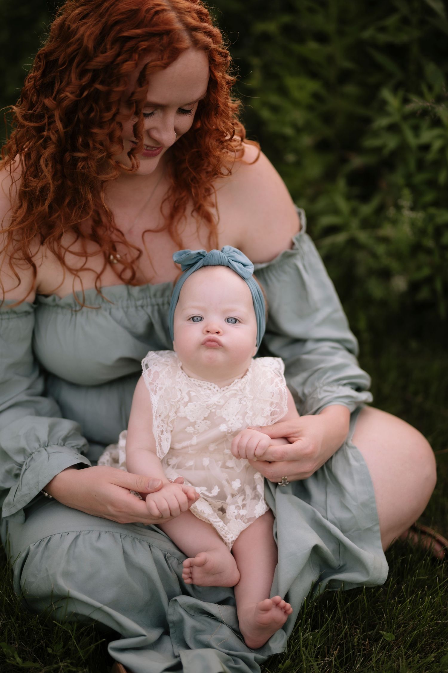 A baby wearing a lace romper and blue headband sits in a parent's lap wearing a sage green dress outdoors.