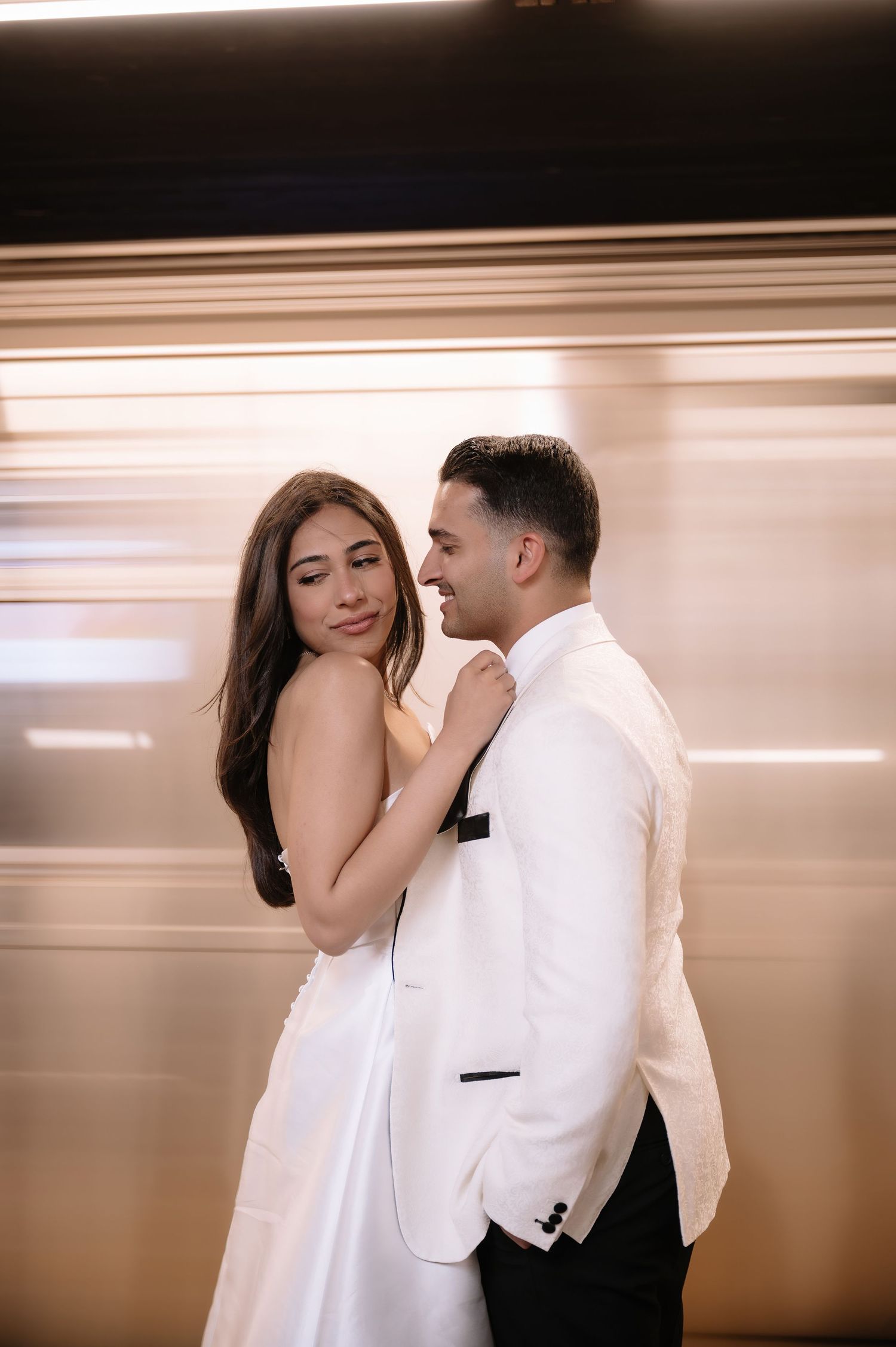 A romantic couple embraces in formal white attire while standing in front of a motion-blurred subway background.