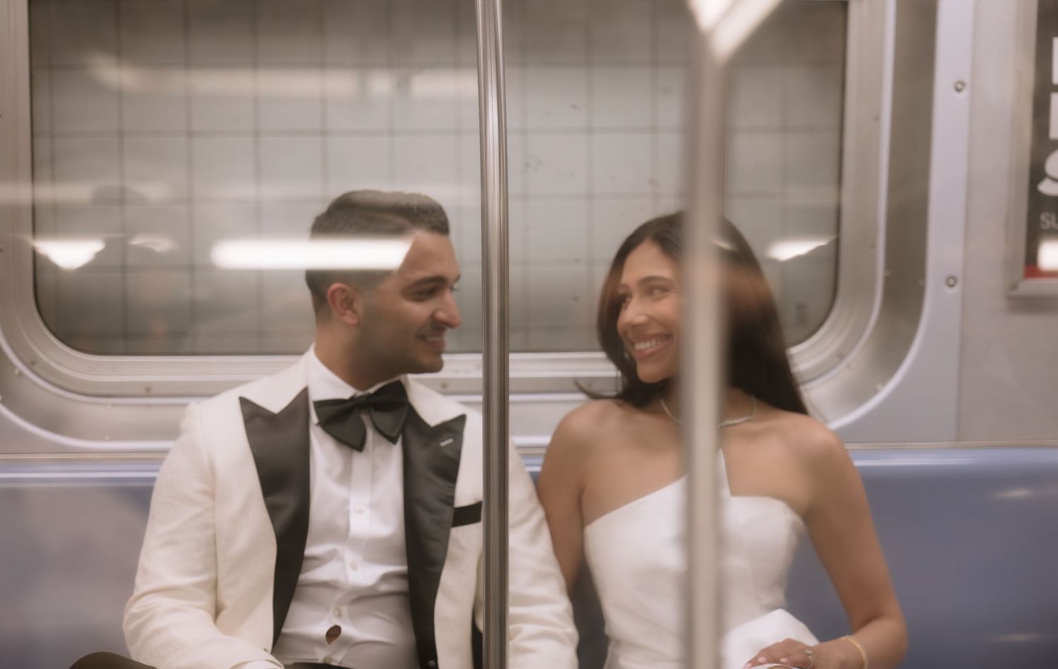 Bride and groom sit together smiling on a subway train during their wedding photoshoot.