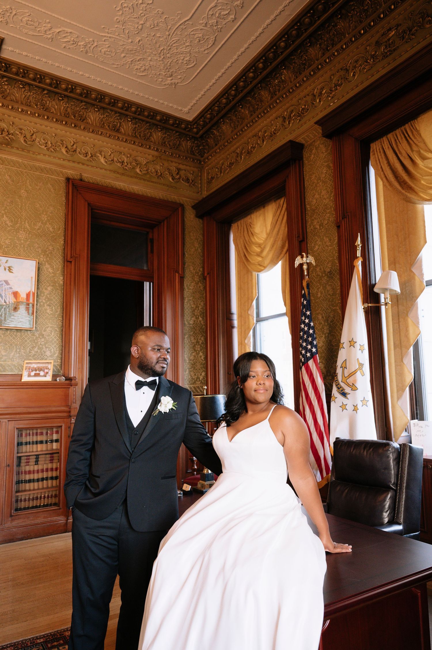 Elegant couple posing in formal wedding attire in classic room with American flag.