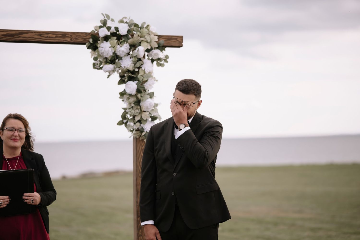 An emotional moment at an outdoor wedding ceremony under a wooden arch decorated with white flowers.