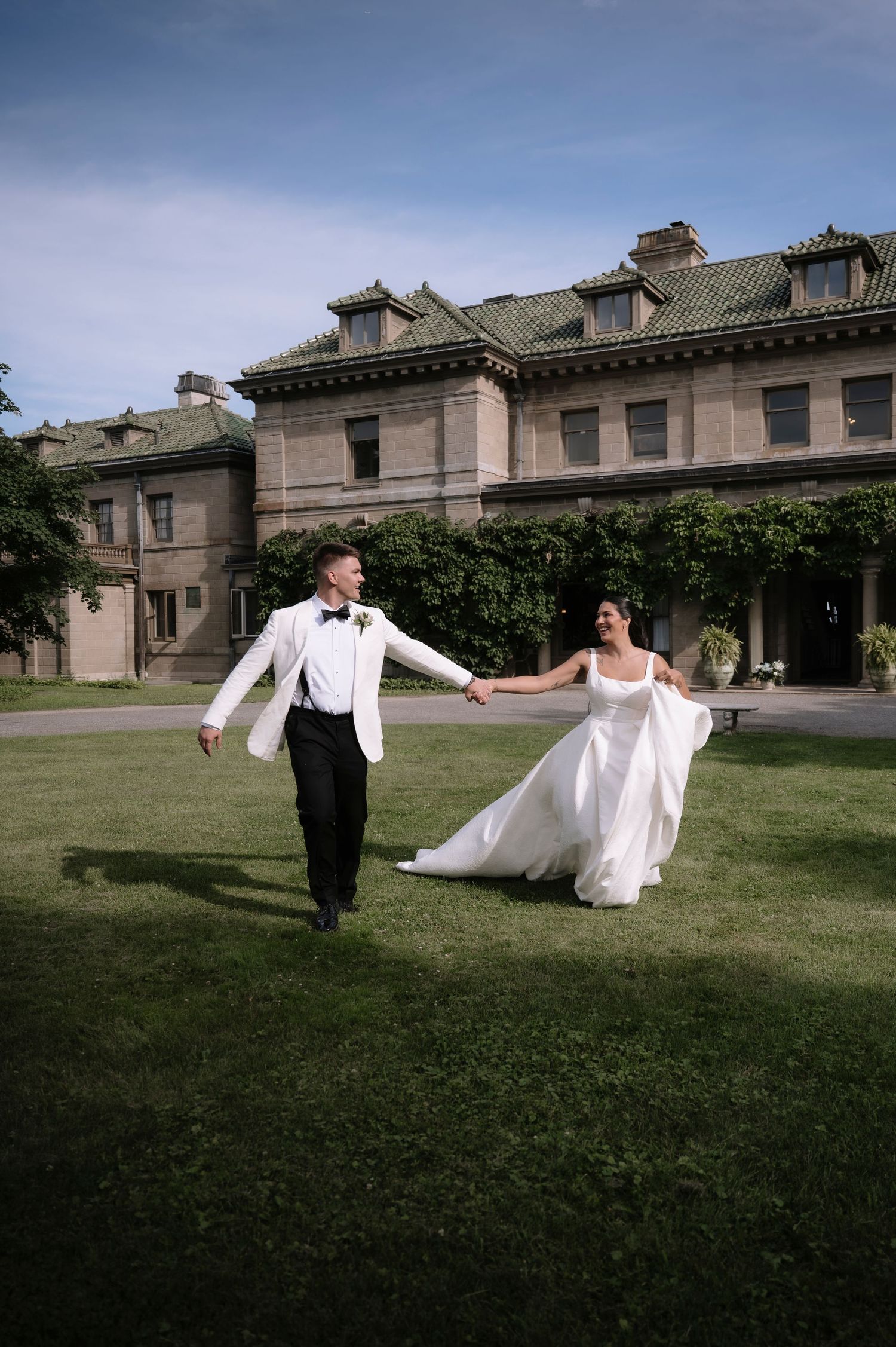 Bride and groom hold hands running across lawn in front of elegant stone mansion.