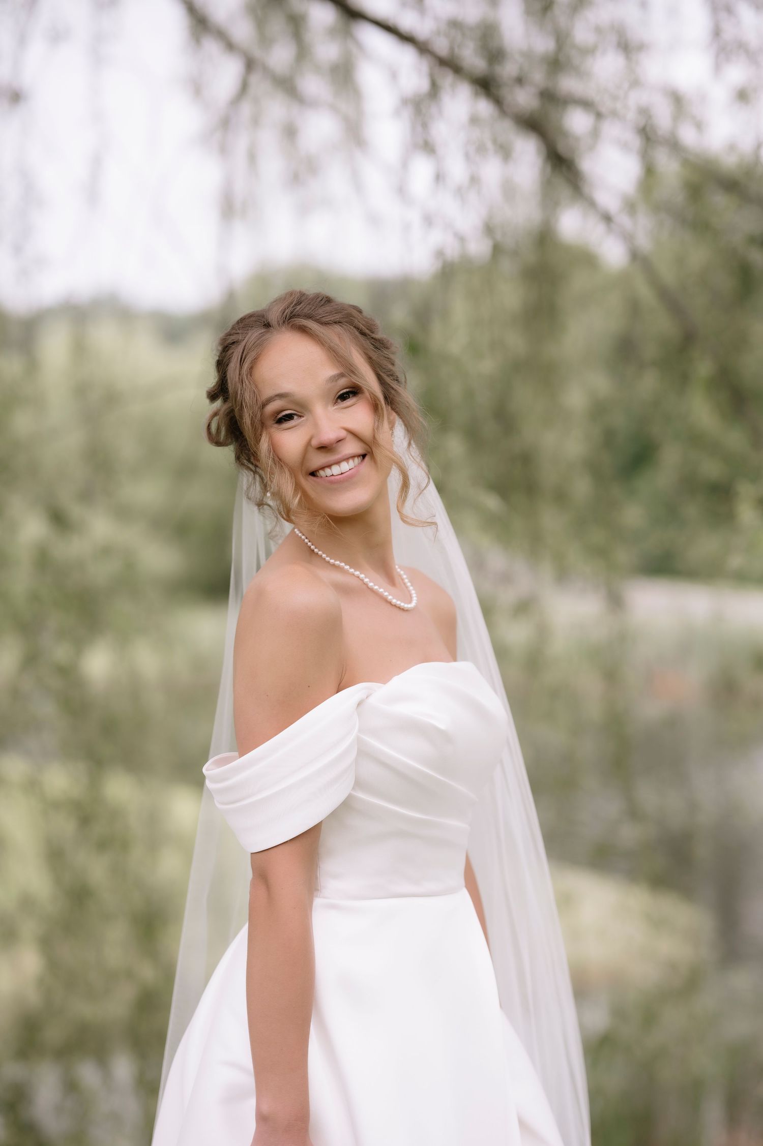 A bride in a classic white off-shoulder wedding dress poses joyfully in an outdoor garden setting with trees in background.