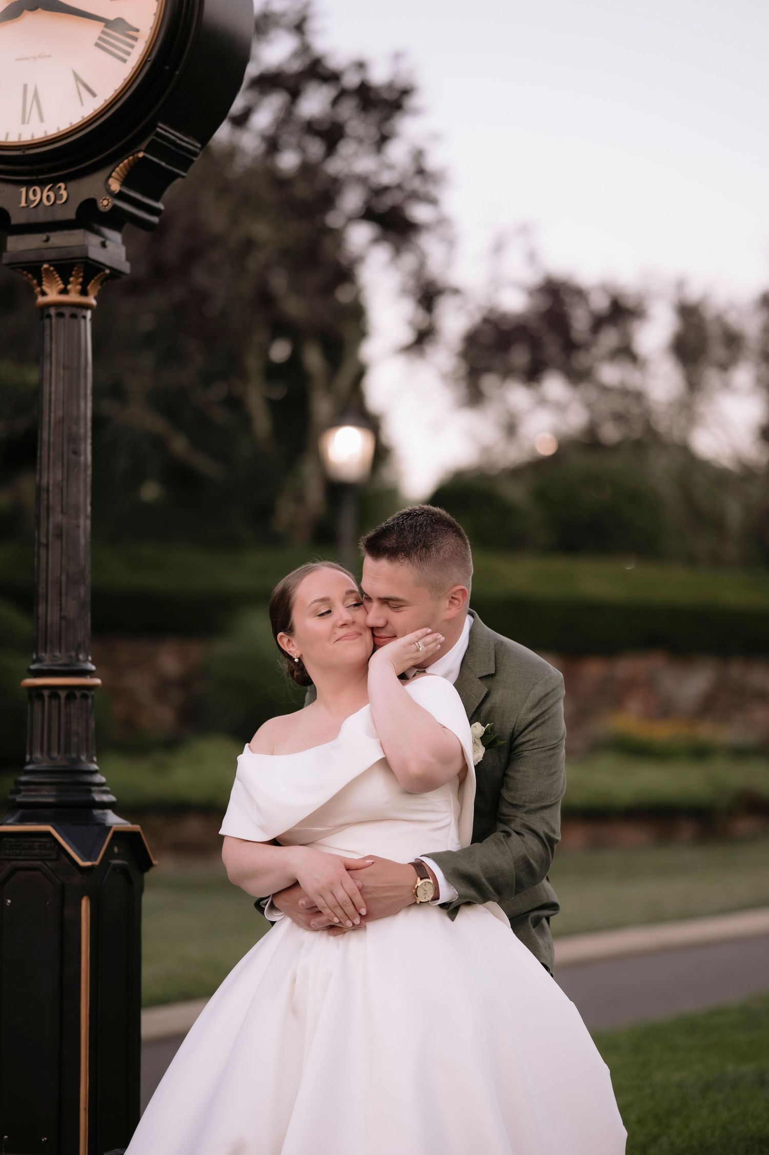 Bride and groom embrace near a vintage 1963 clock at dusk, groom in green suit kissing bride's cheek.