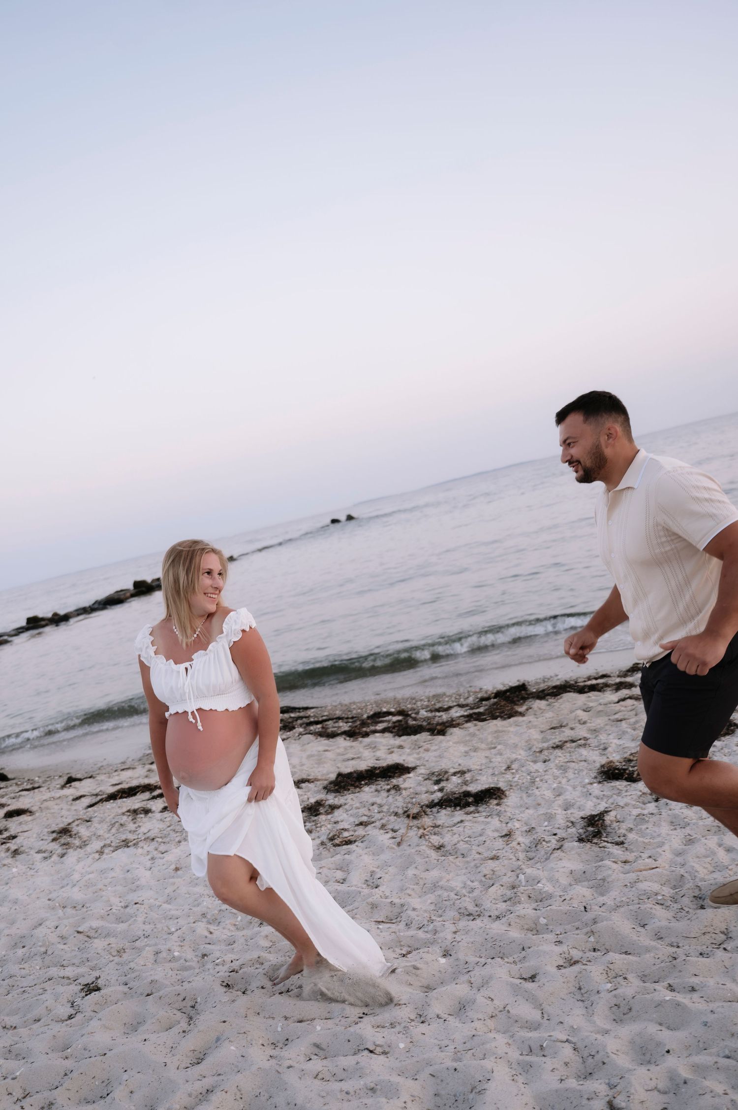 Pregnant woman in white dress laughing as man runs toward her on sandy beach at sunset.