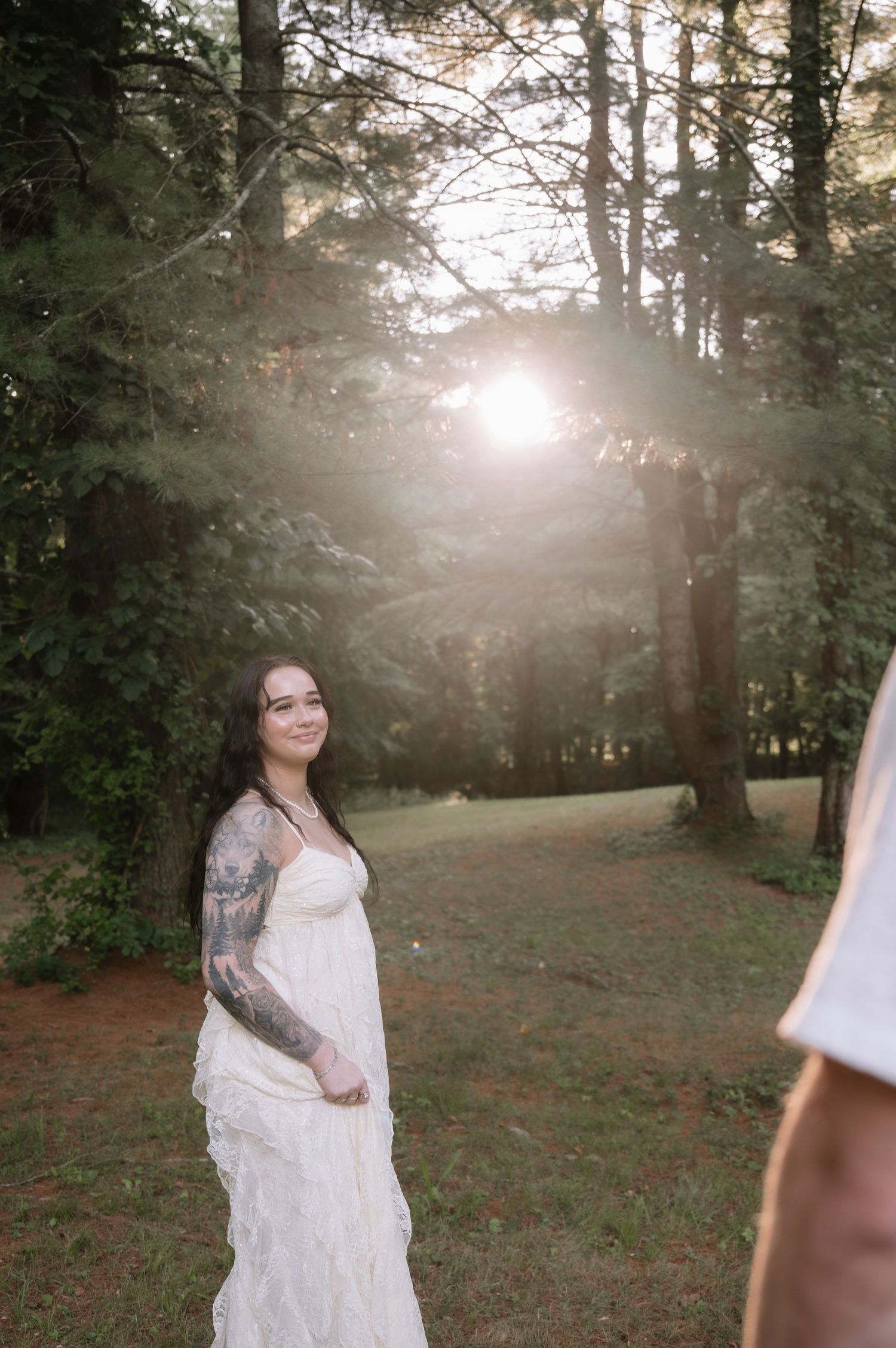 Tattooed bride in white lace gown smiling in sunlit forest setting during golden hour outdoor wedding.