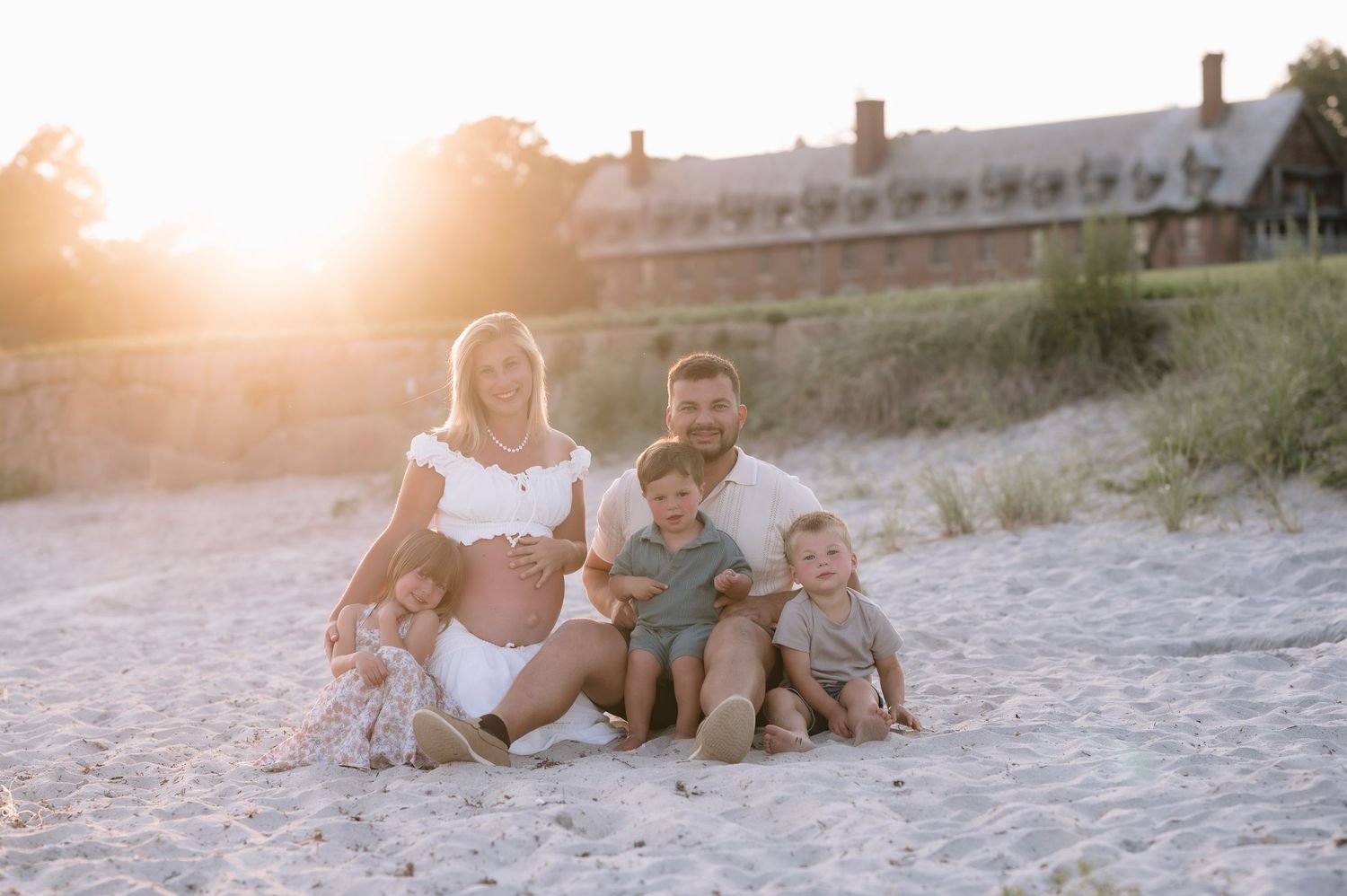 Pregnant woman and man sit on sandy beach at sunset with three young children in front of a large building.