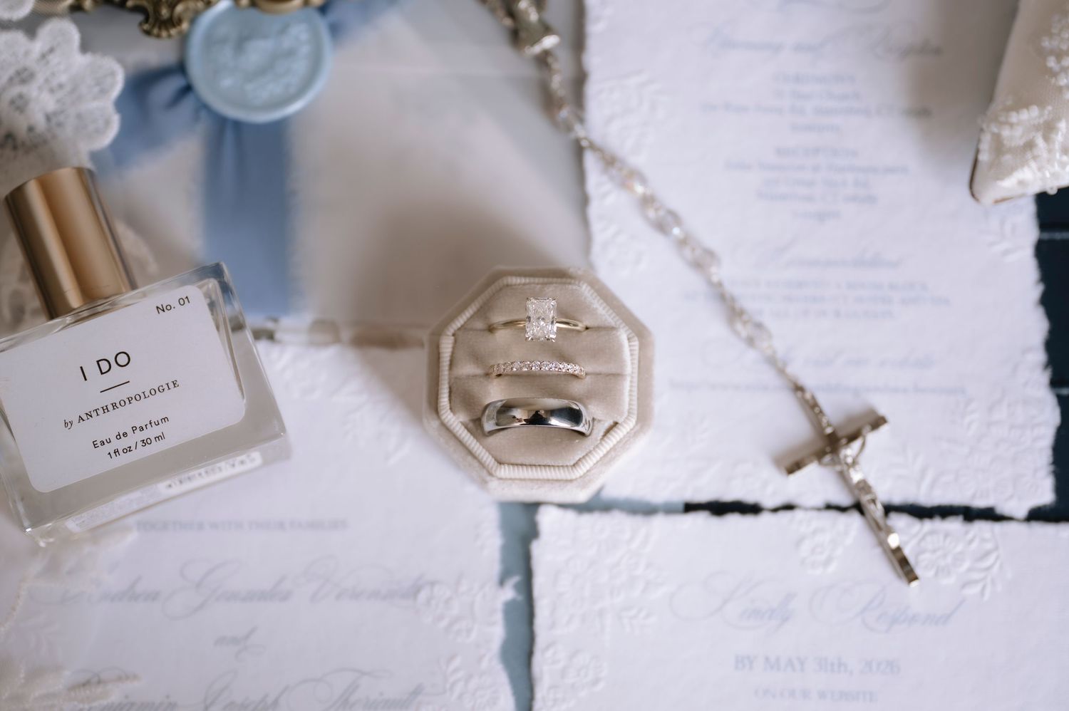 Elegant wedding flat lay with ring box, diamond rings, rosary, perfume bottle, and blue ribbon on embossed stationery.