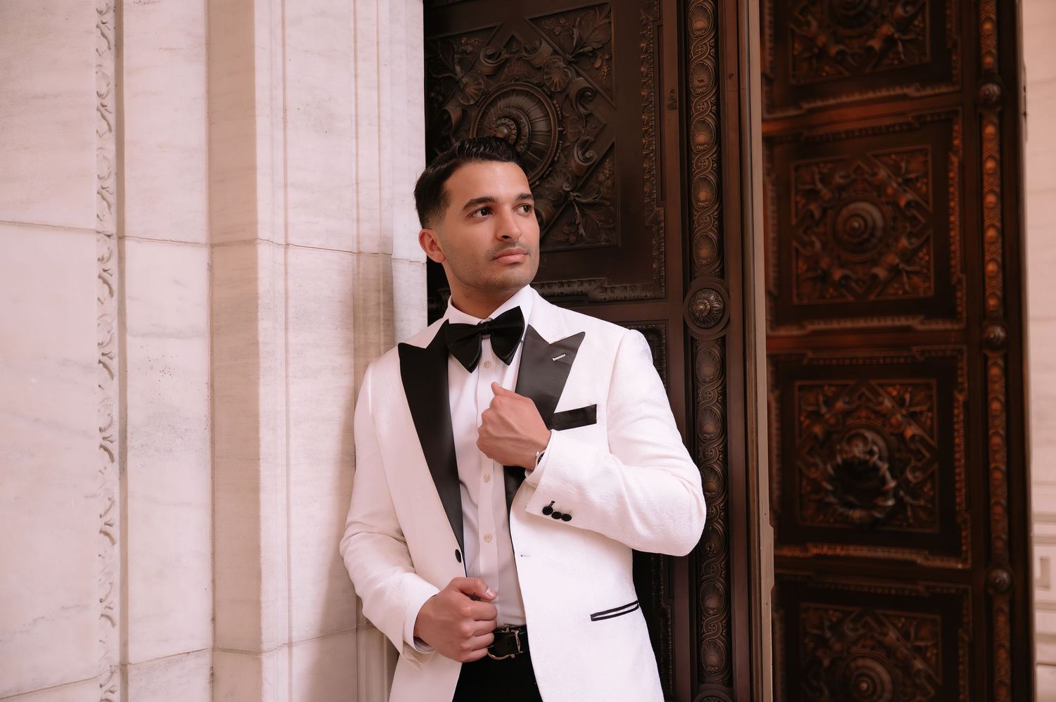 A person wearing a classic white tuxedo jacket with black bow tie poses elegantly against an ornate wooden door.