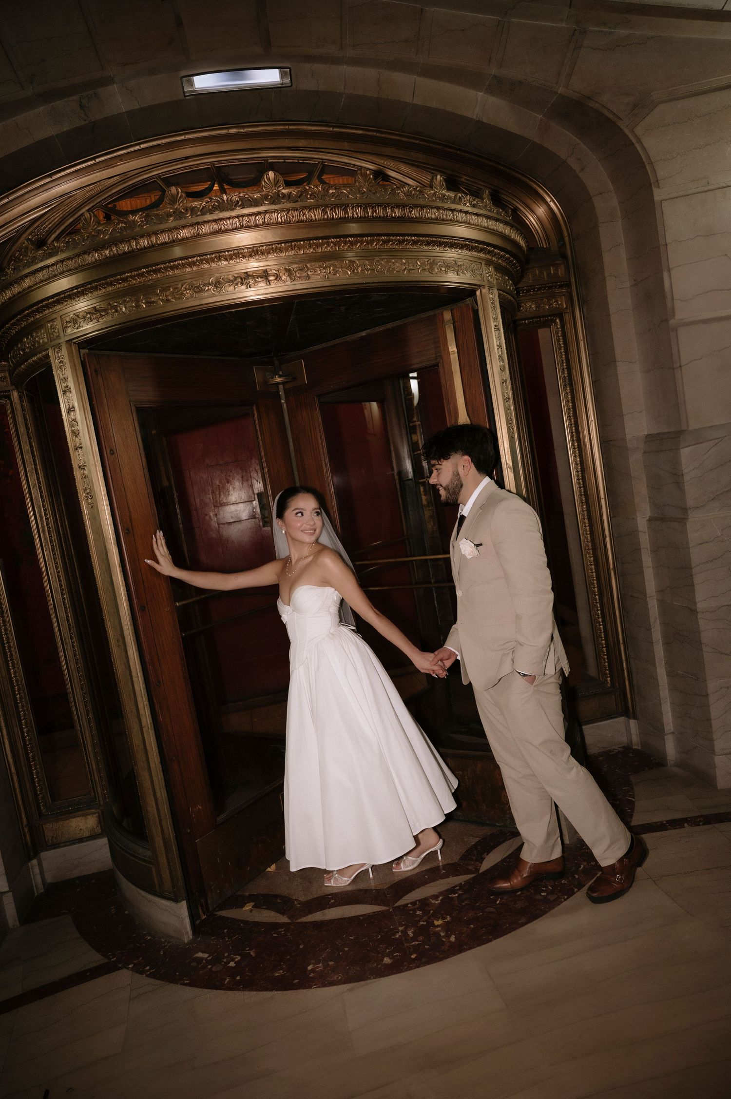 A couple in wedding attire playfully poses in a dark vintage hallway with ornate architectural details.