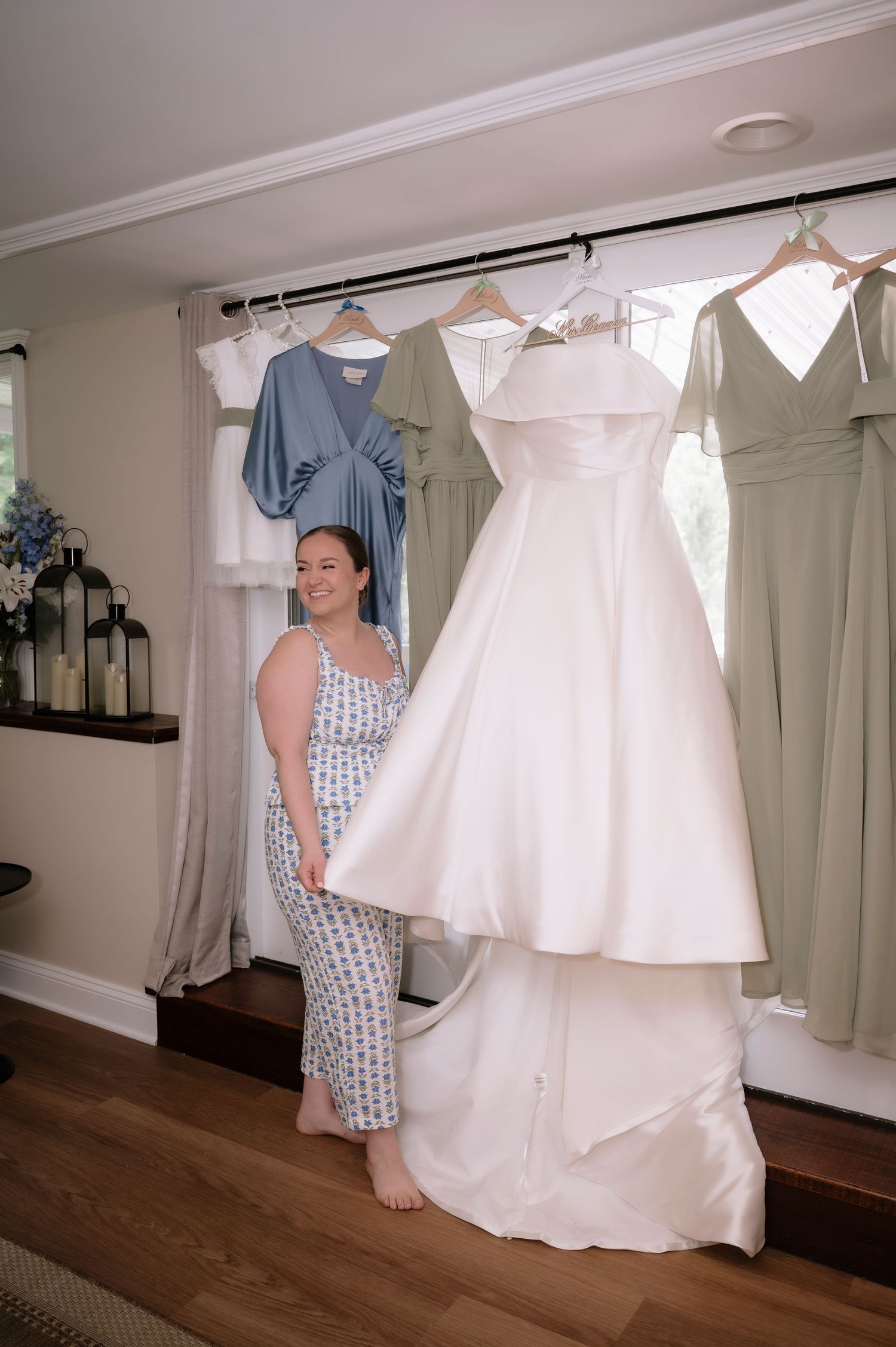 Bride in pajamas smiling beside her elegant white wedding gown hanging with colorful bridesmaid dresses in a bright room.