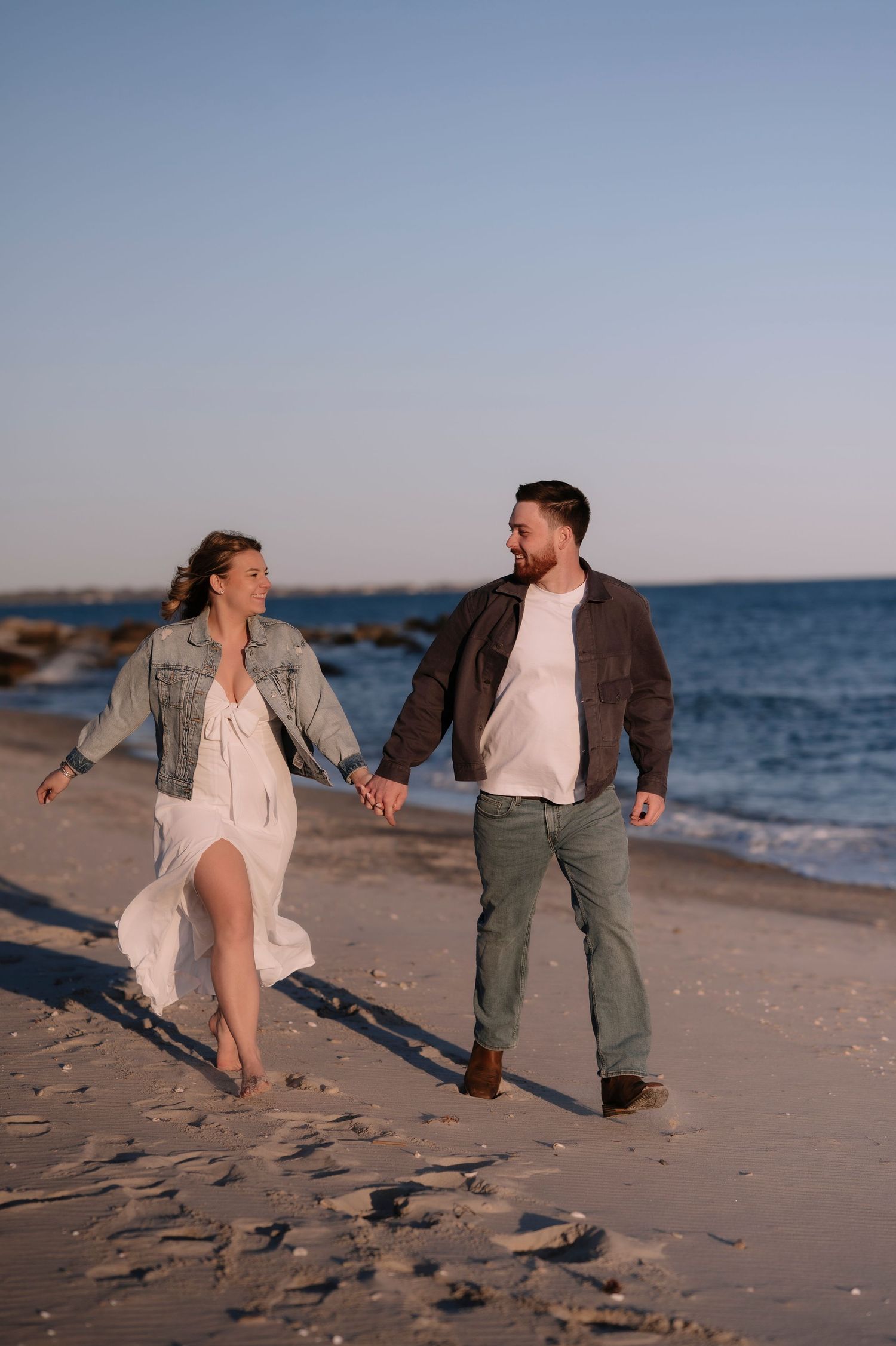 Couple holding hands and walking on a sandy beach at sunset, woman in white dress and denim jacket, man in brown jacket.