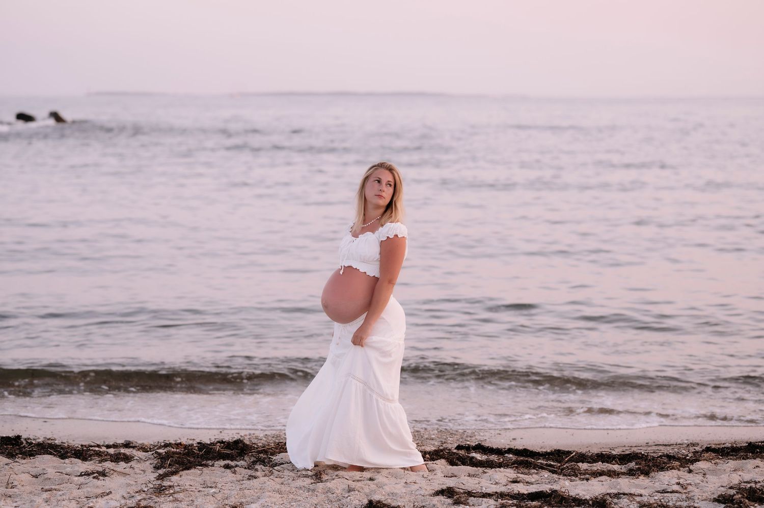 Pregnant woman in white dress cradling baby bump on sandy beach at sunset with calm ocean waves behind her.