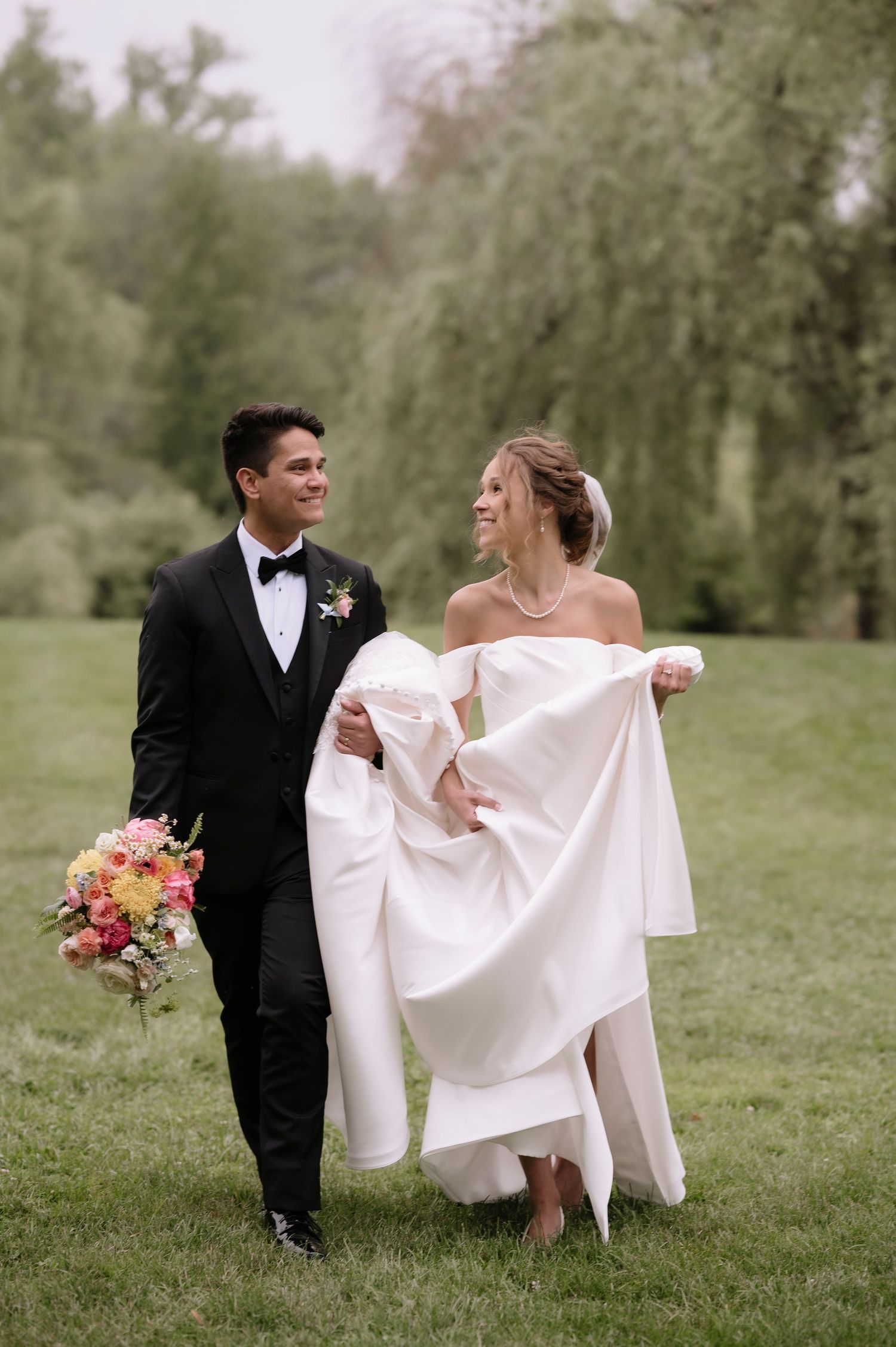 Newlywed couple holding hands and walking through a grassy field with white dress and black tuxedo in spring.