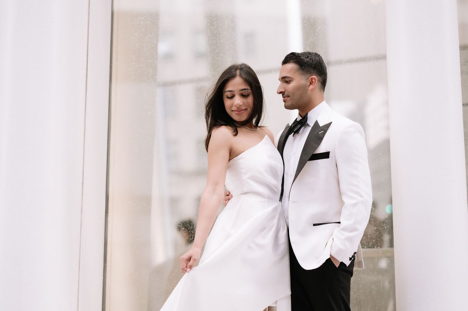 A couple in elegant white formal wear pose against a modern architectural backdrop.