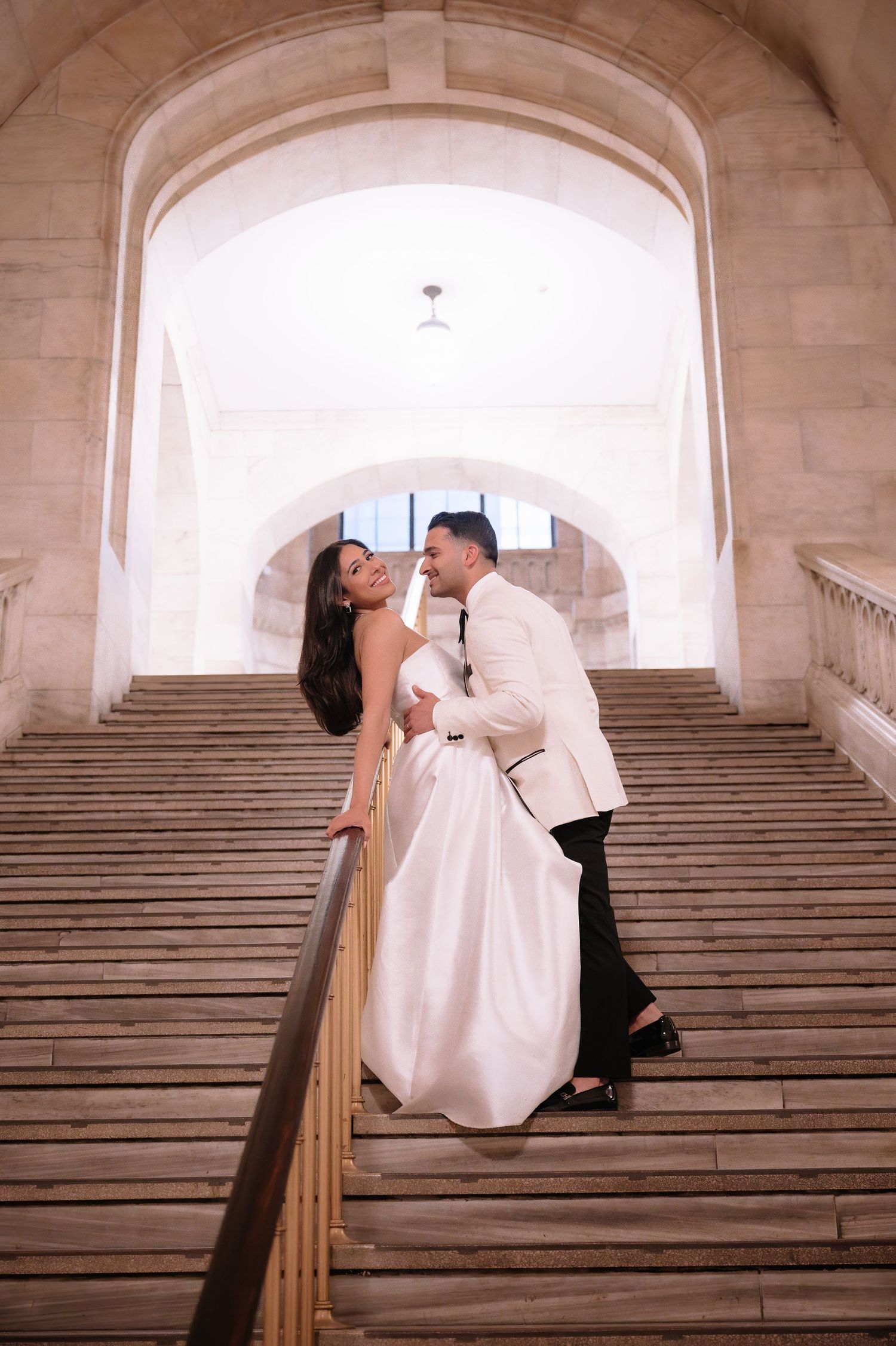A romantic couple in wedding attire share an intimate moment on grand stone steps under an arched passageway.