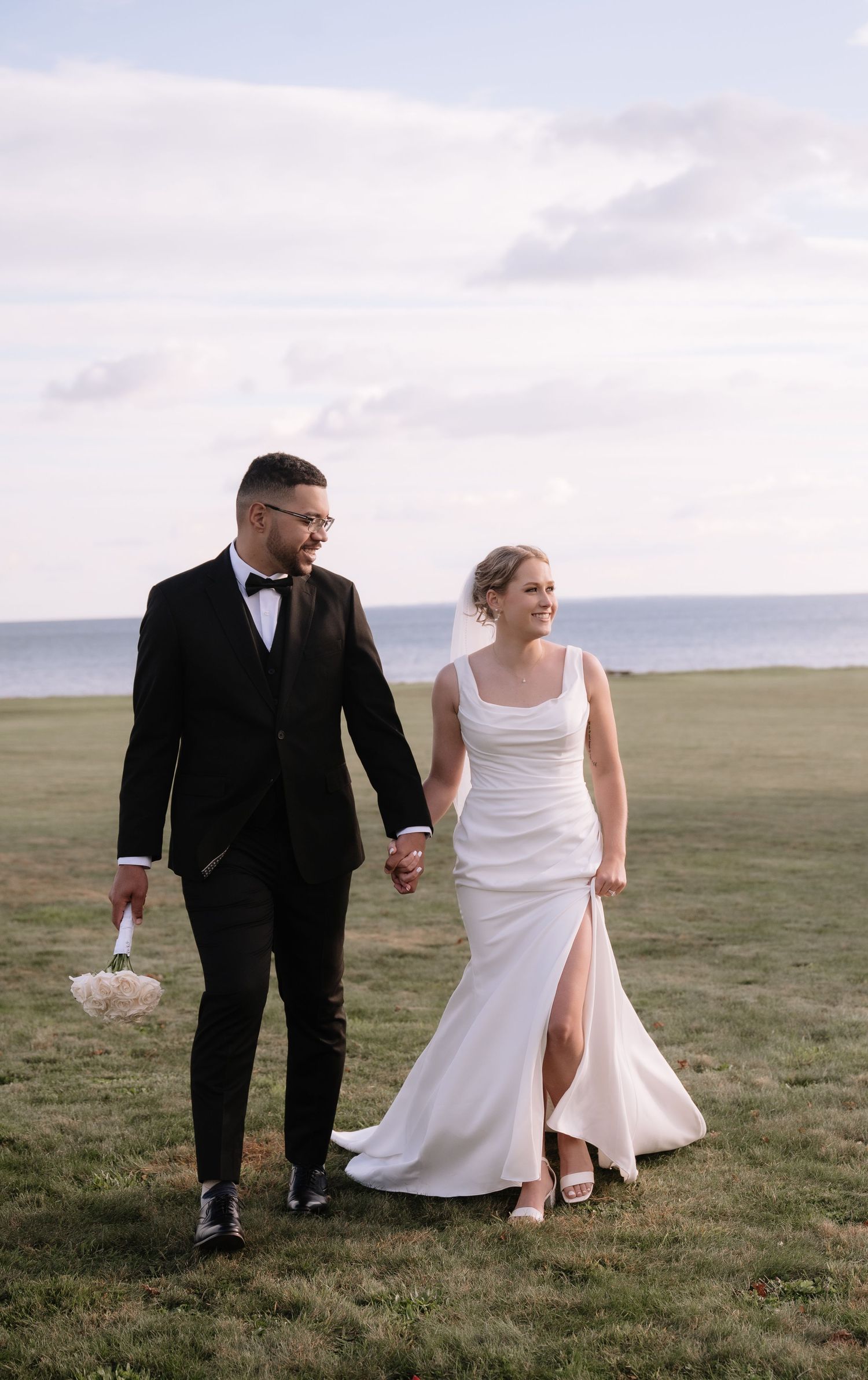 A couple in wedding attire walks together across a scenic green hillside against a cloudy sky.