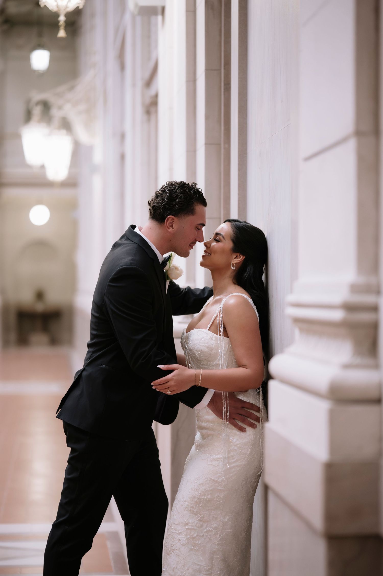 Bride in lace gown and groom in black suit share an intimate moment leaning against elegant marble columns.
