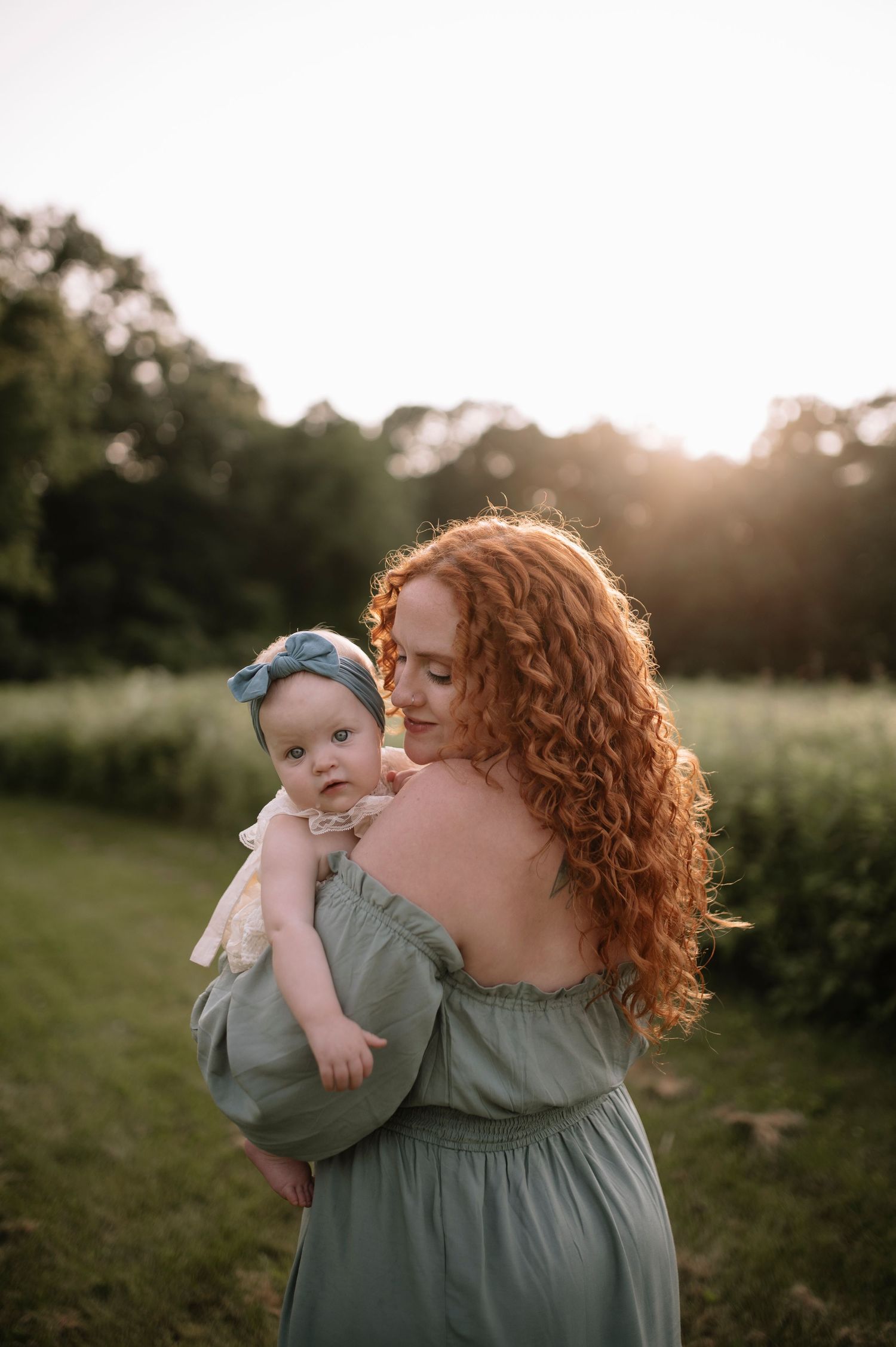 Mother in sage green dress holds baby outdoors at sunset while standing in grassy field.