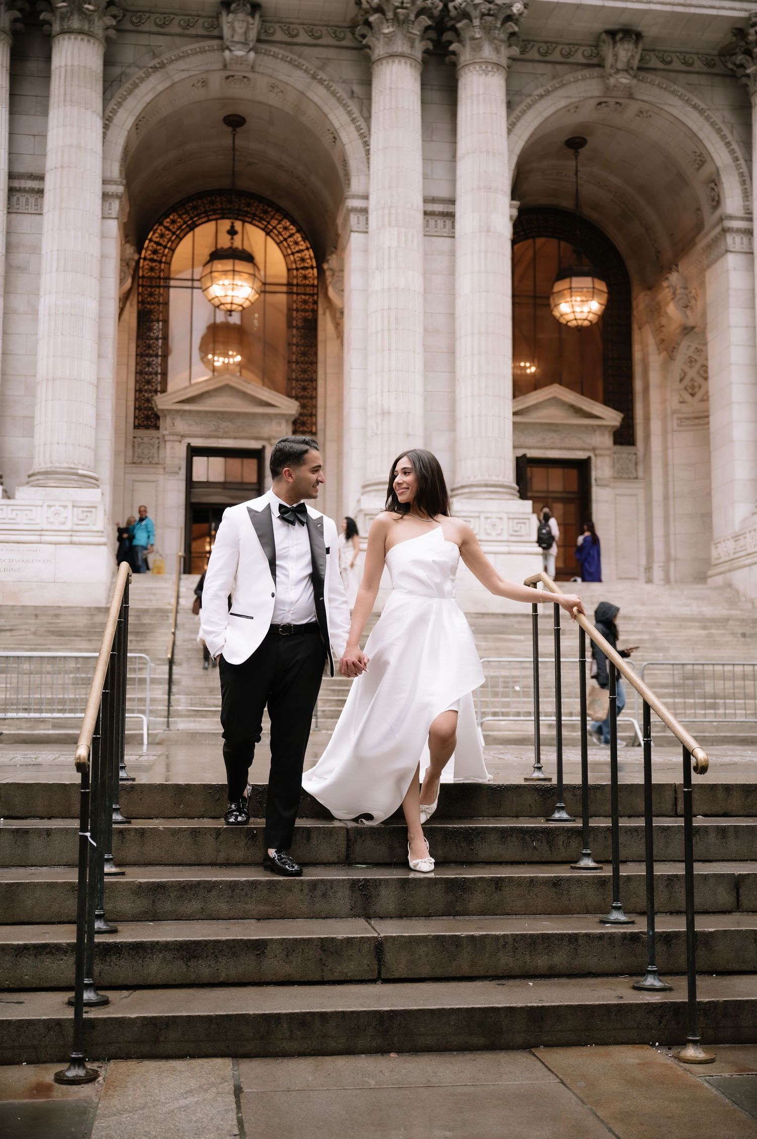 Elegant newlyweds descending ornate stone steps of classical building.
