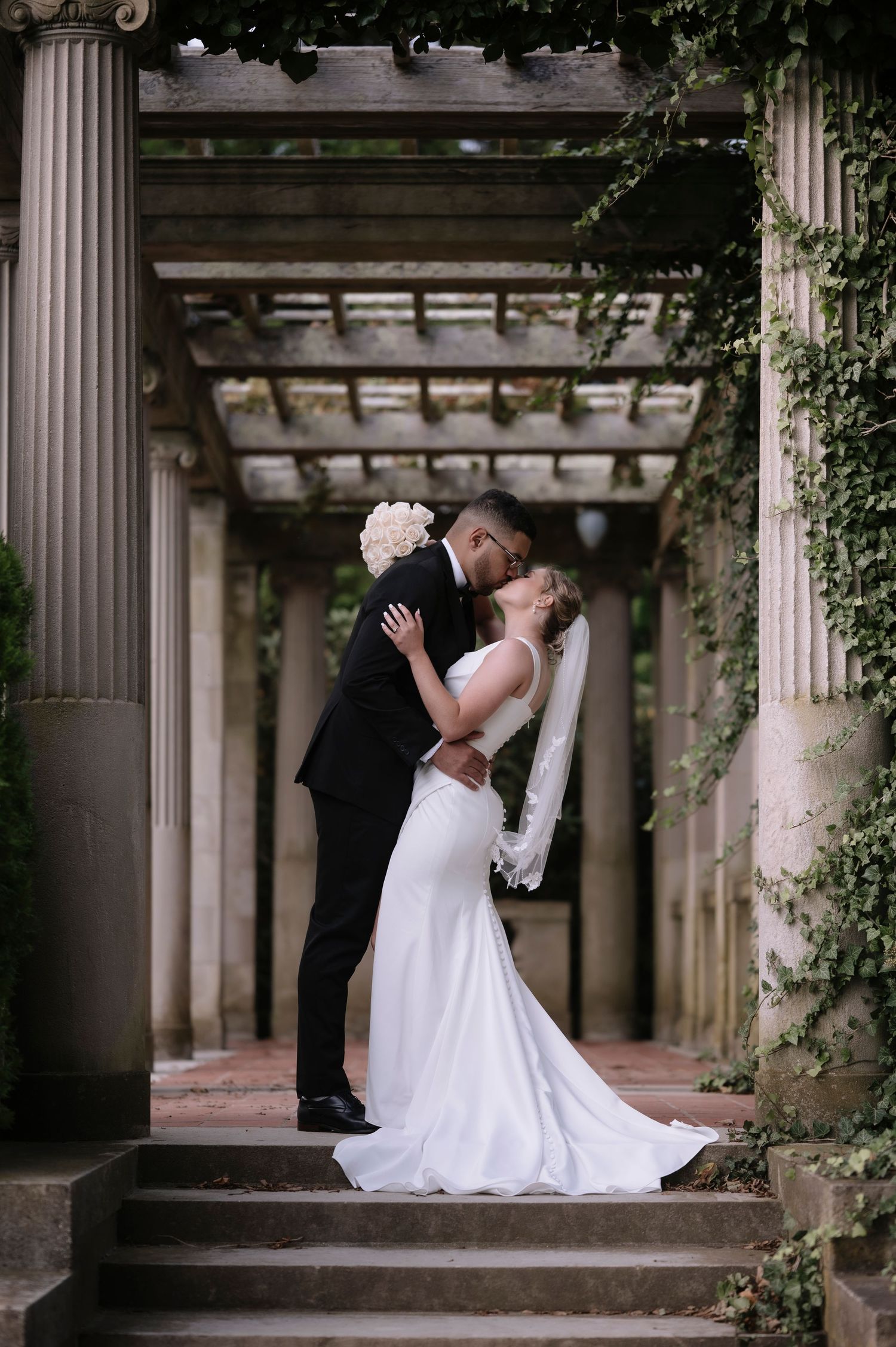 A romantic wedding couple embraces under an elegant columned pergola with climbing vines.