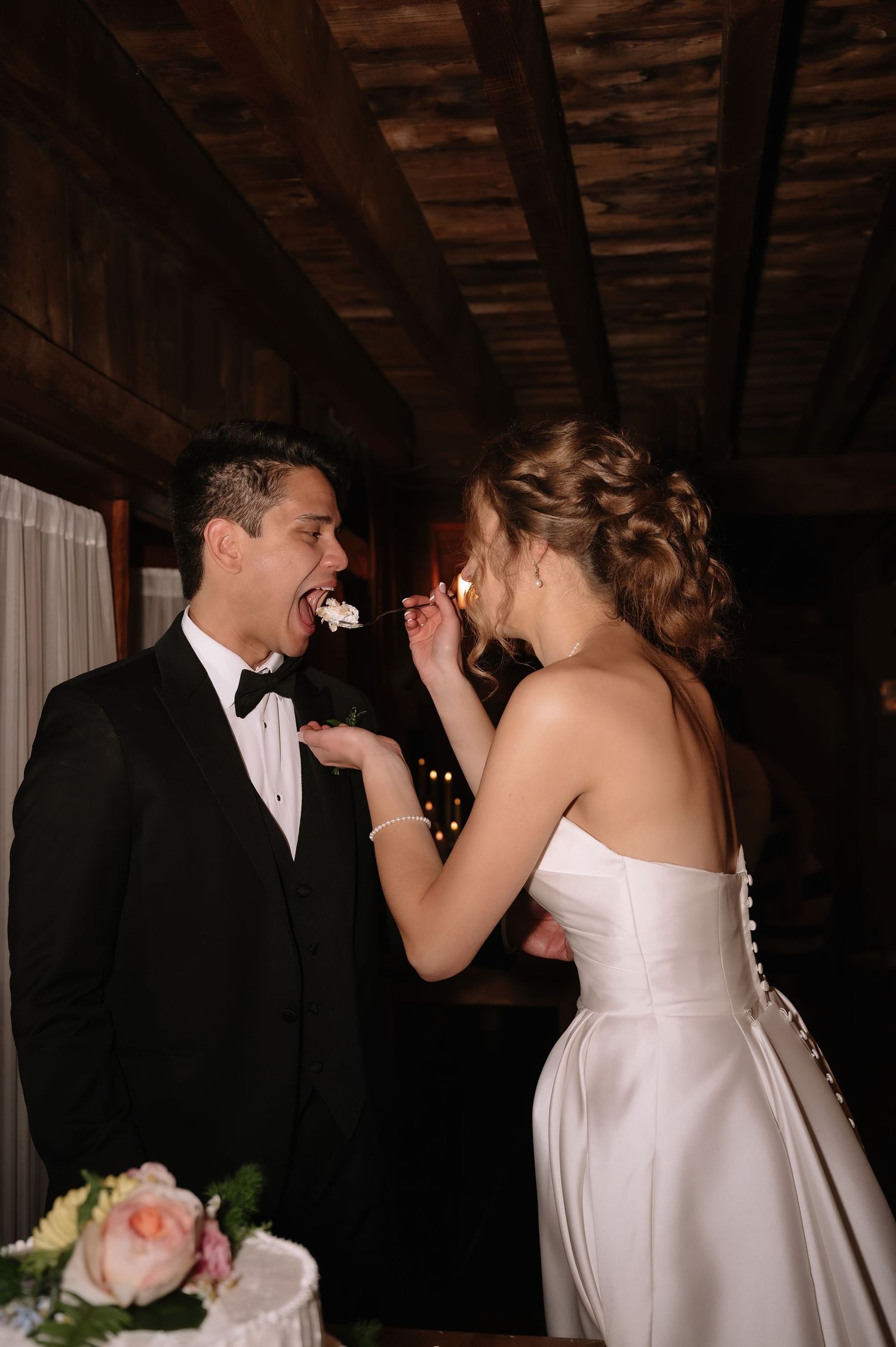 Bride in strapless white gown feeds wedding cake to groom in black tuxedo at rustic indoor reception.