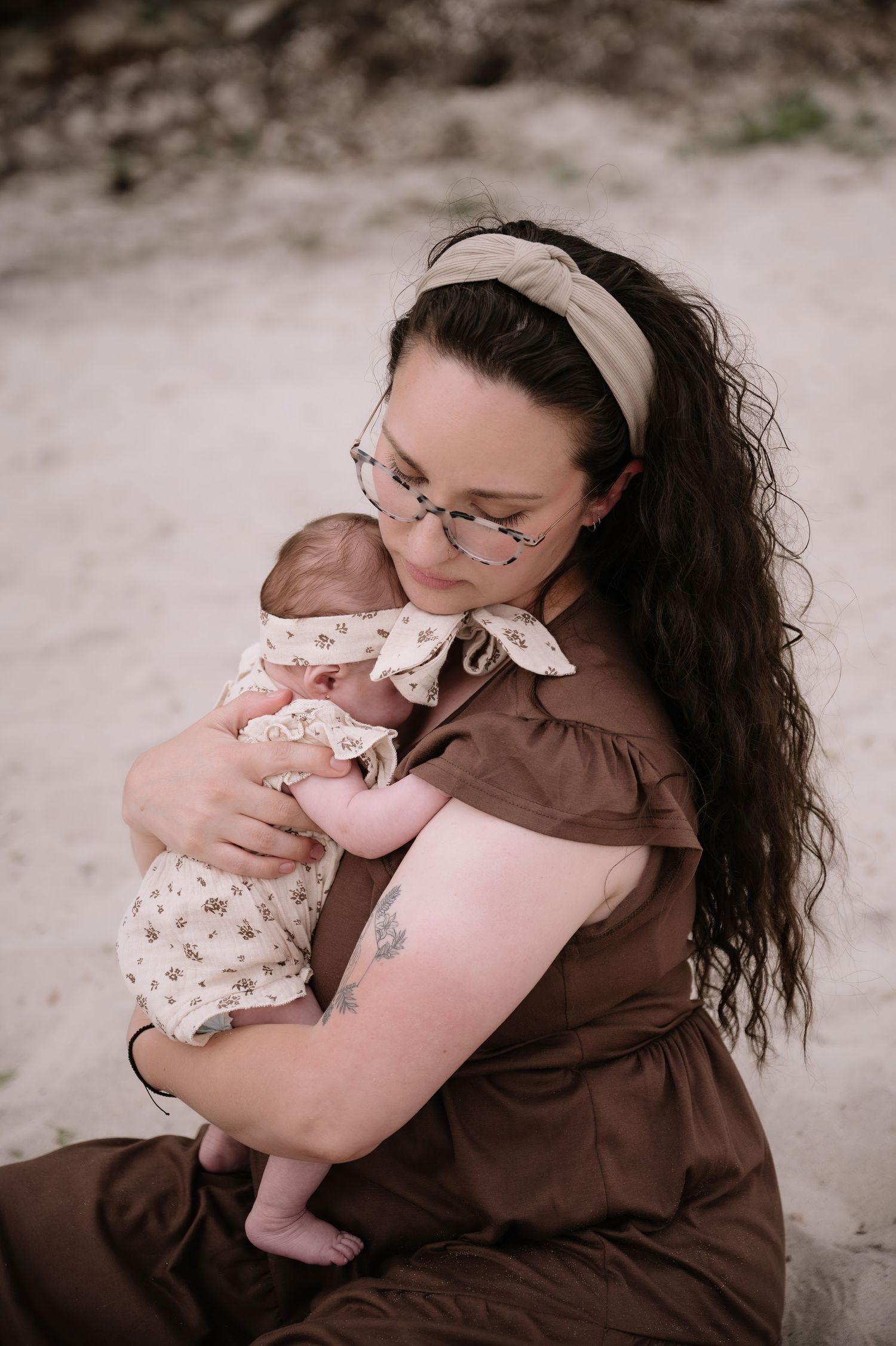 A mother in a brown dress holding an infant wearing floral clothing sits outdoors in a candid moment.