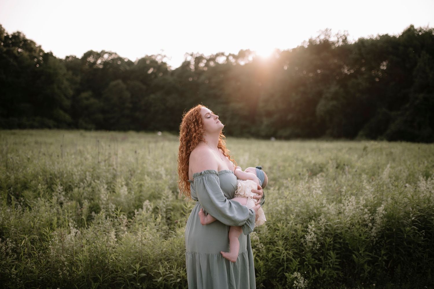 A figure stands in a sunlit meadow wearing a flowing sage green dress while holding a baby at sunset.