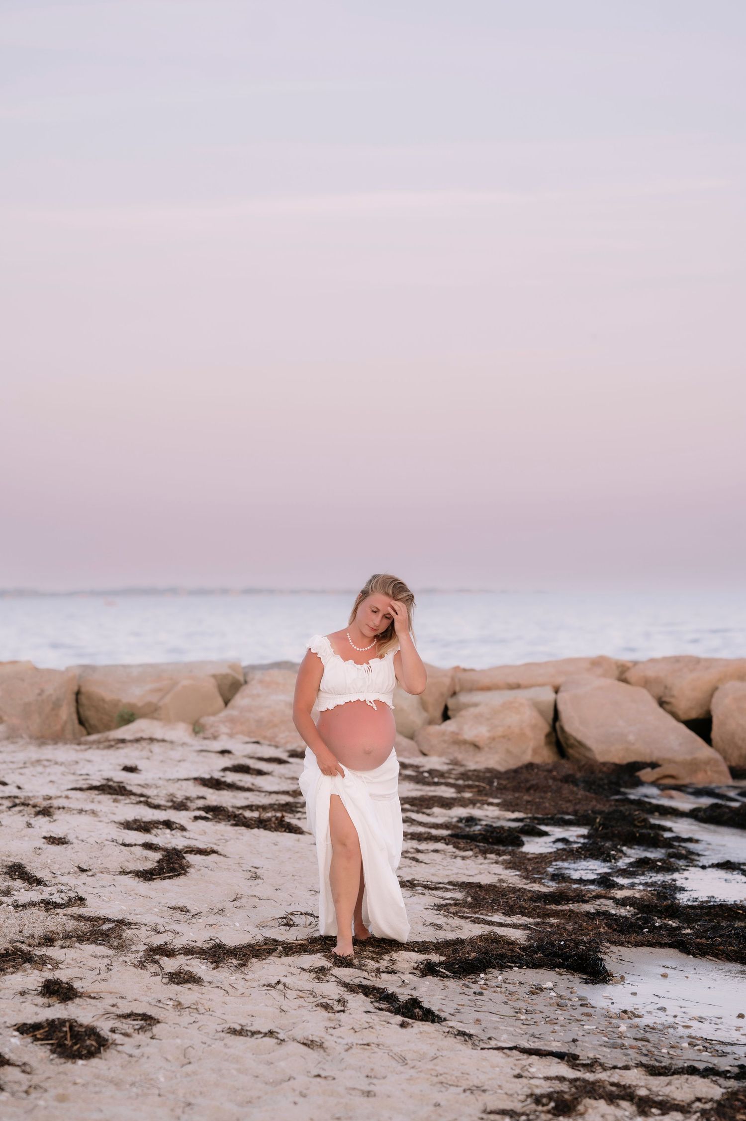 Pregnant woman in white crop top and skirt standing on a sandy beach at sunset with rocky shoreline backdrop.