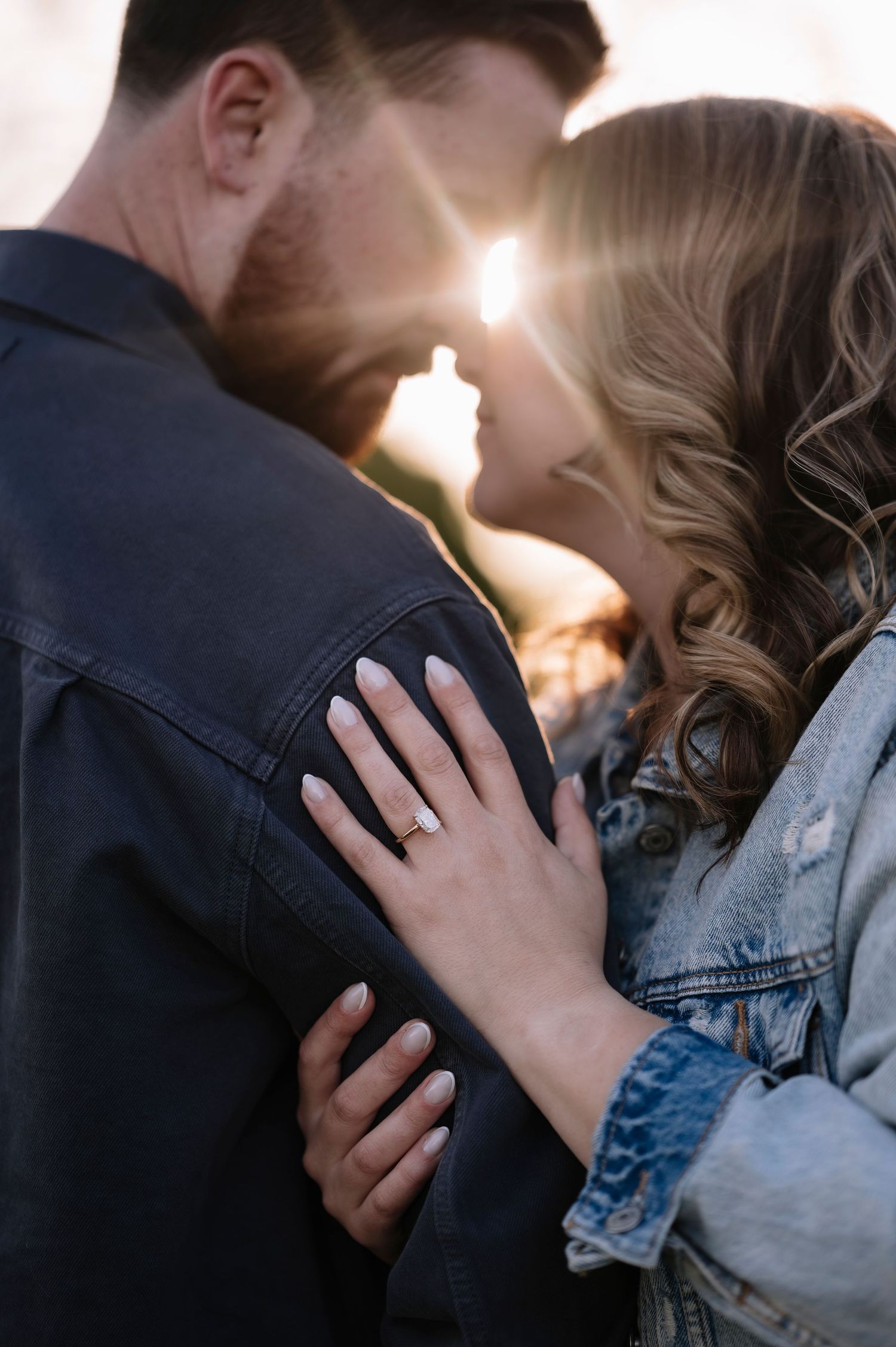 Close-up of engaged couple nearly kissing at sunset, woman's hand showing diamond ring on man's dark jacket.