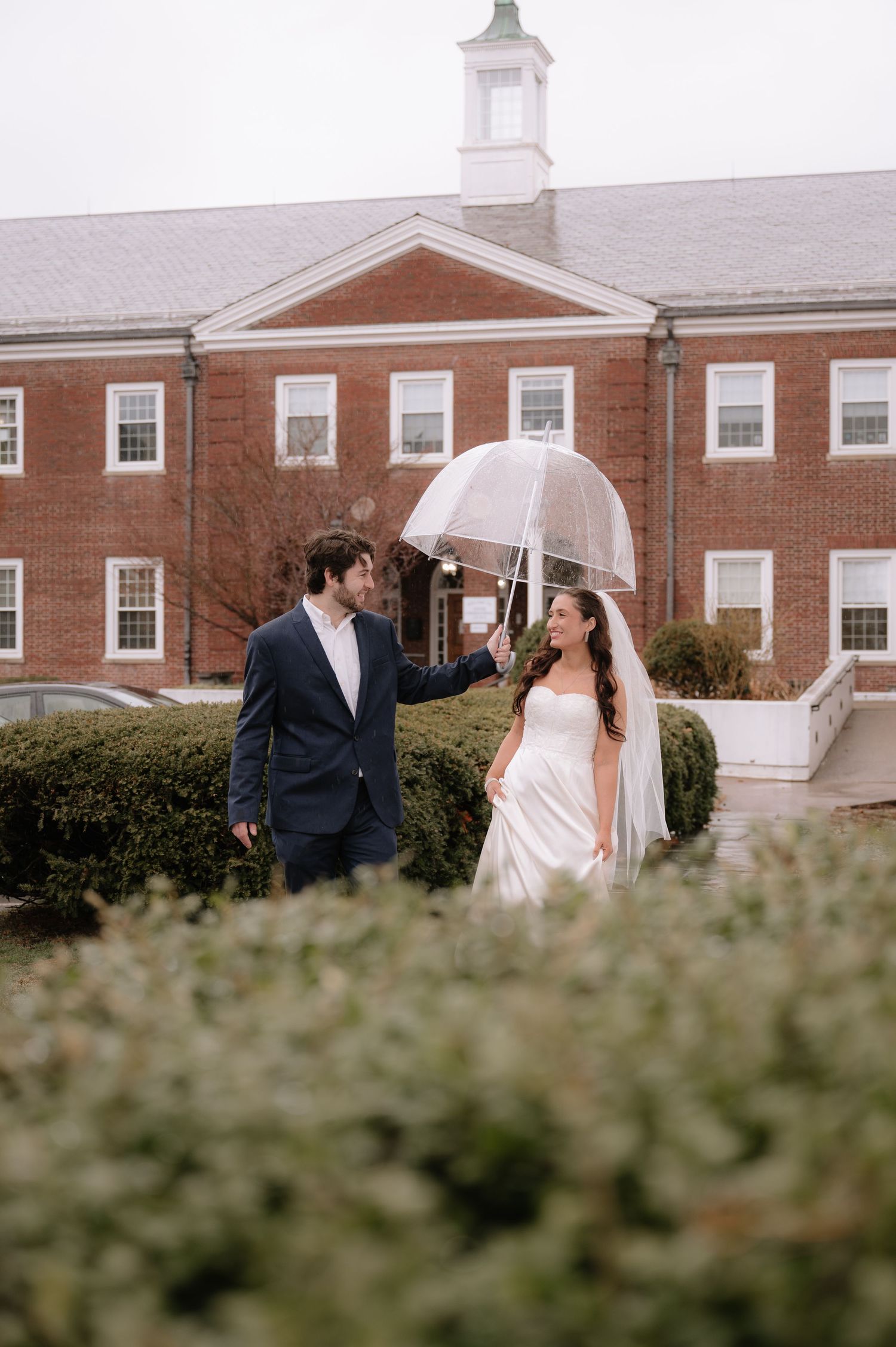 Couple walking in front of a brick building as groom holds umbrella over bride.