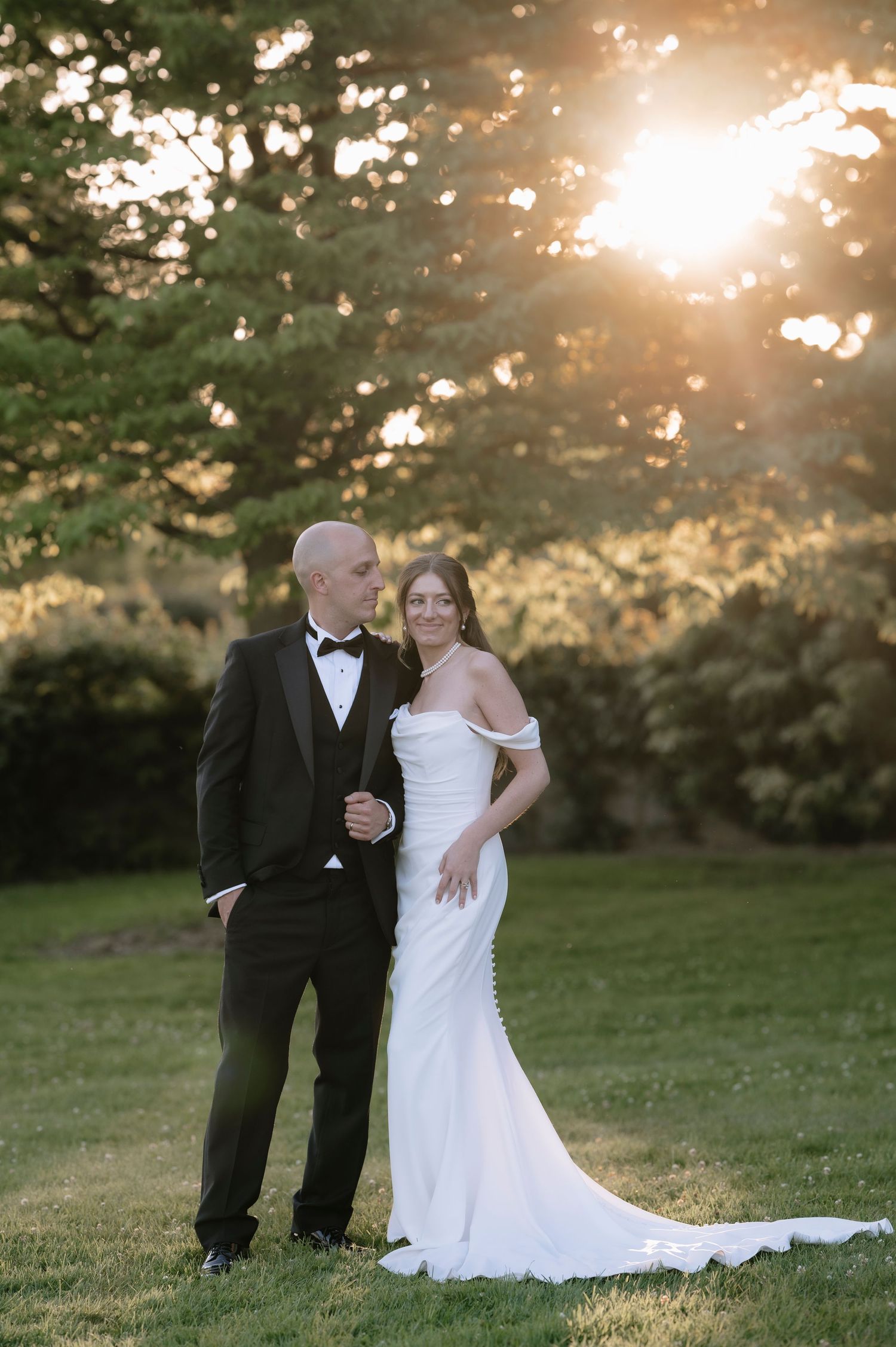 A couple in wedding attire embraces during golden hour sunset in a grassy field with trees in the background.