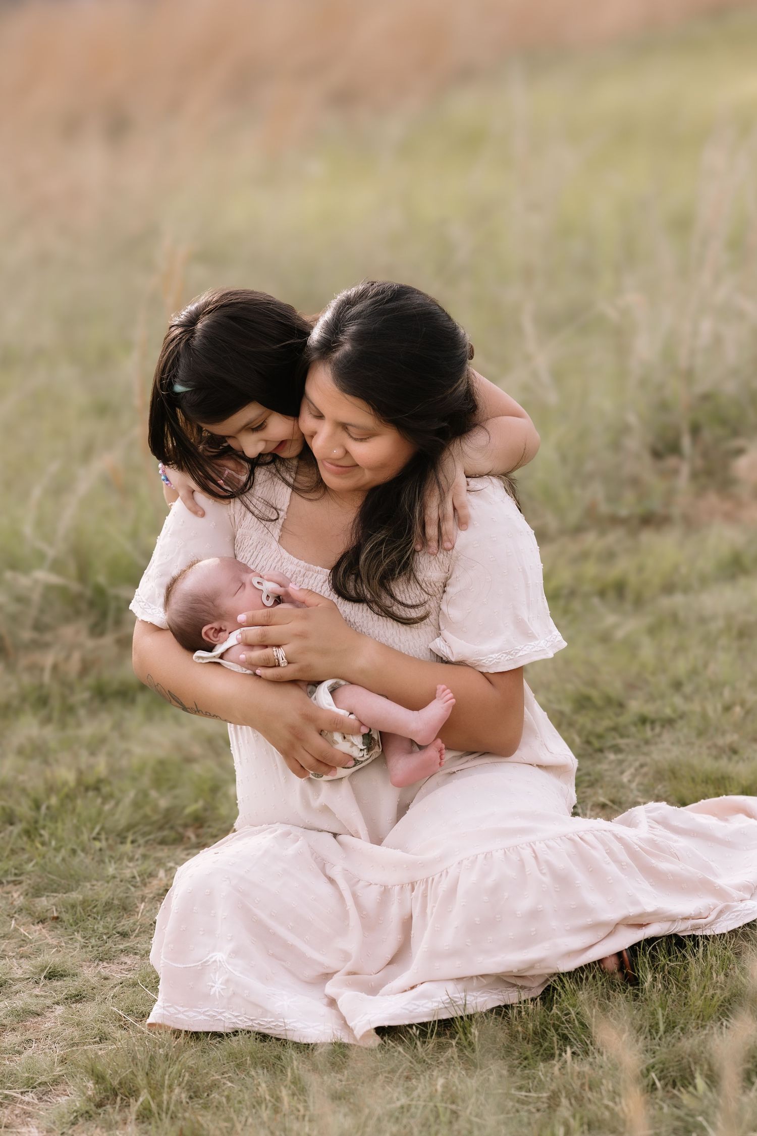 A mother sits in a field holding her newborn while her young daughter embraces her from behind, all in pink.