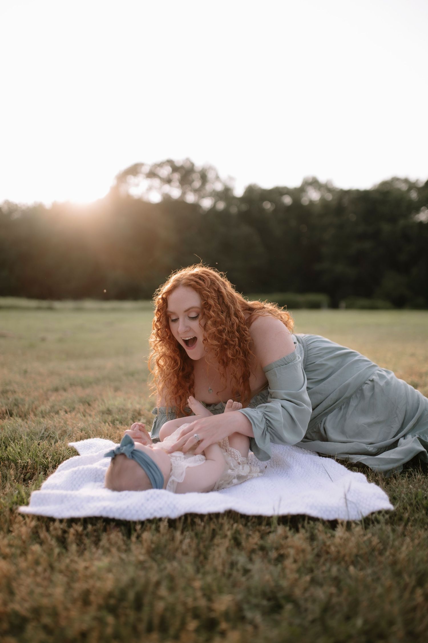 A tender sunset moment captured on a blanket in a grassy field, with glowing warm light and curly ginger hair in focus.