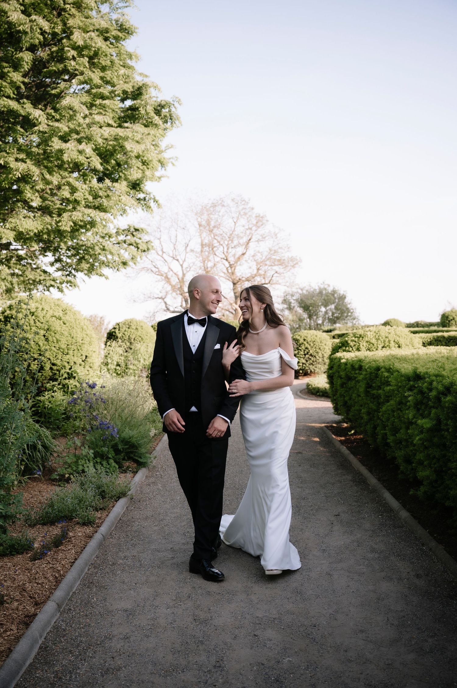 A bride and companion share a joyful moment walking down a tree-lined garden path during an outdoor wedding ceremony.