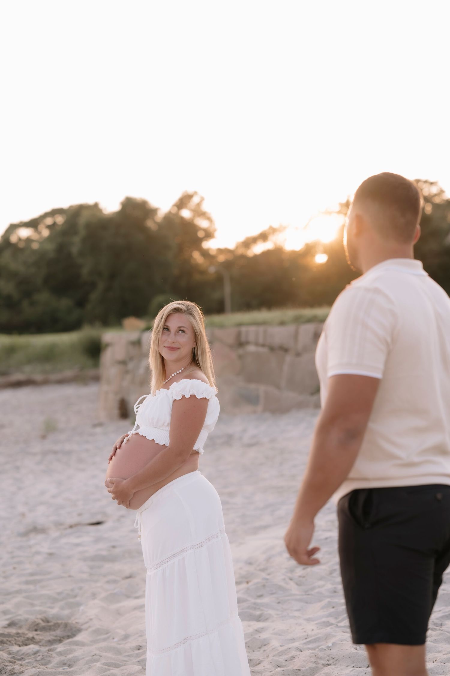 Pregnant woman in white off-shoulder dress holds baby bump on sandy beach at golden sunset with partner nearby.