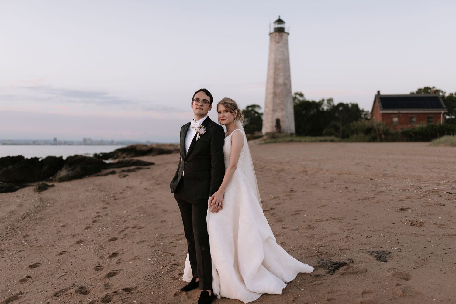 Bride and groom posing on sandy beach near historic lighthouse at dusk during their wedding day.