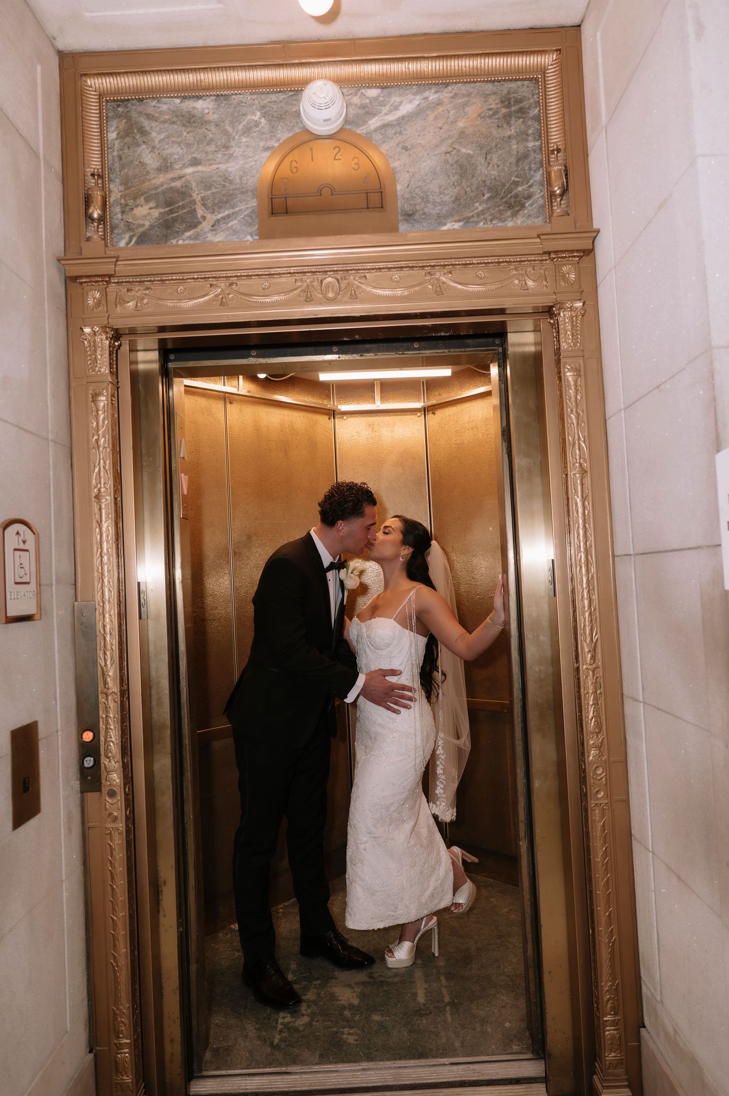 Bride and groom sharing a romantic kiss inside an ornate gold elevator on their wedding day.
