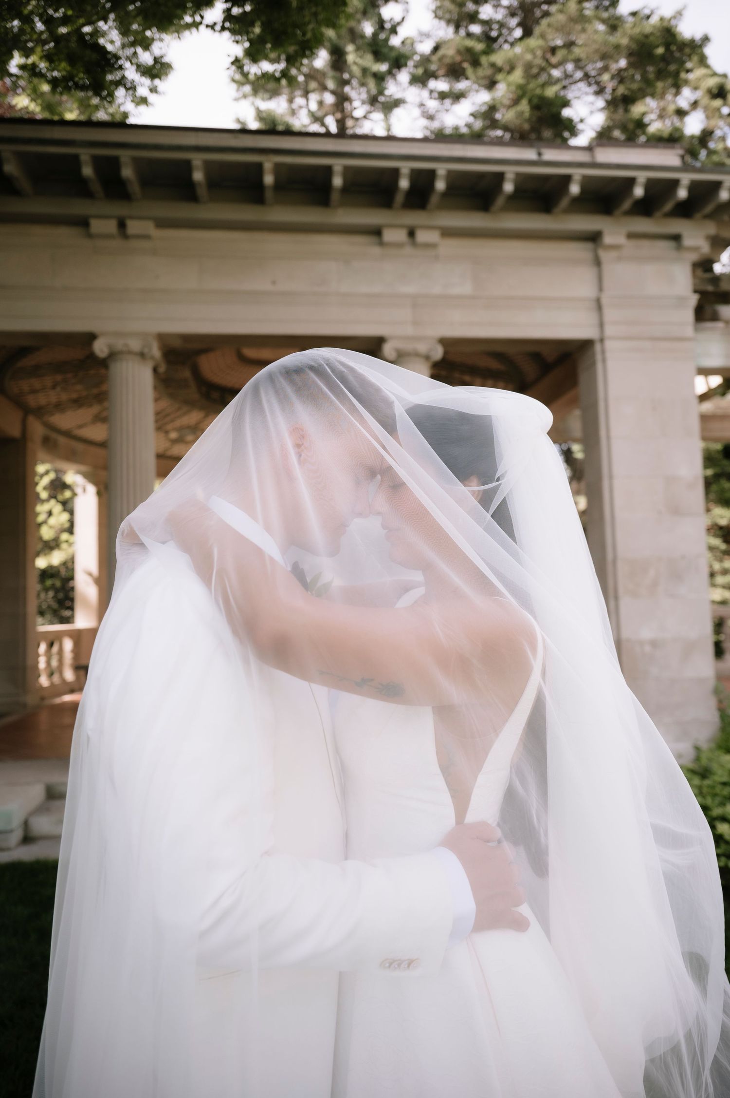 Bride and groom share a romantic kiss hidden beneath a flowing white wedding veil outdoors.
