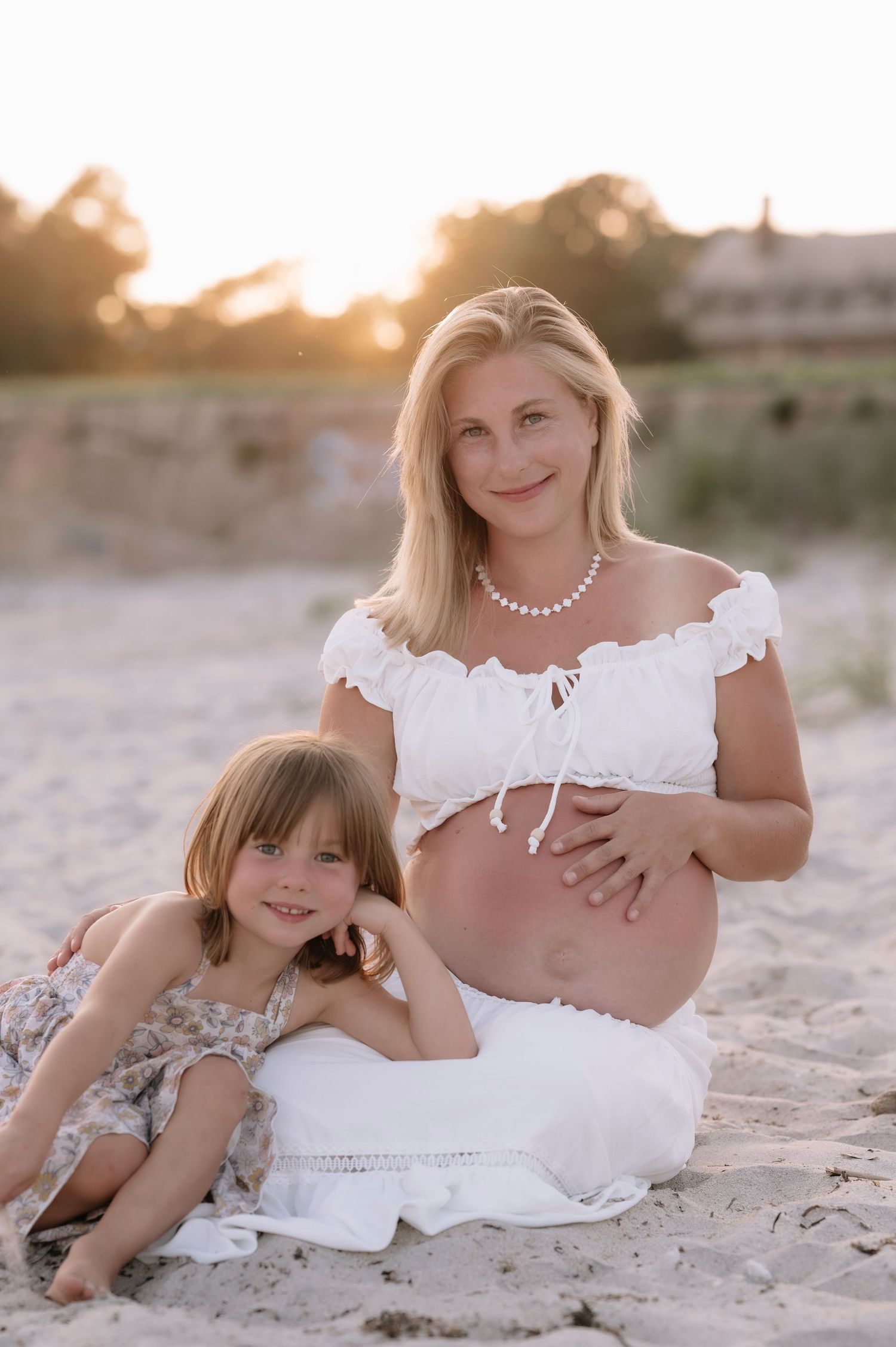 Pregnant woman in white crop top and skirt sits on beach with young daughter at golden hour sunset.