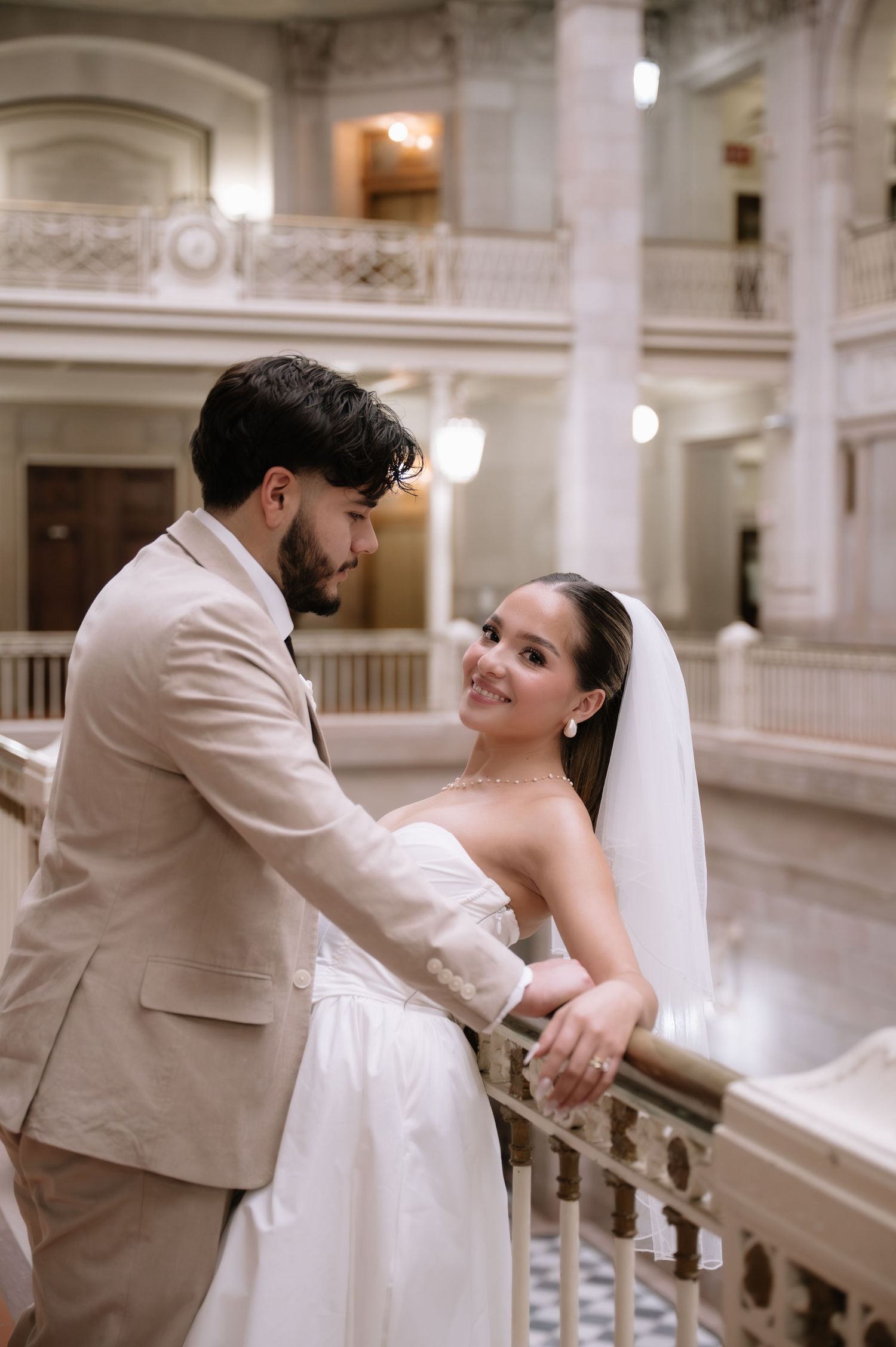 A romantic wedding couple dances together in an elegant white marble interior venue with ornate architectural details.